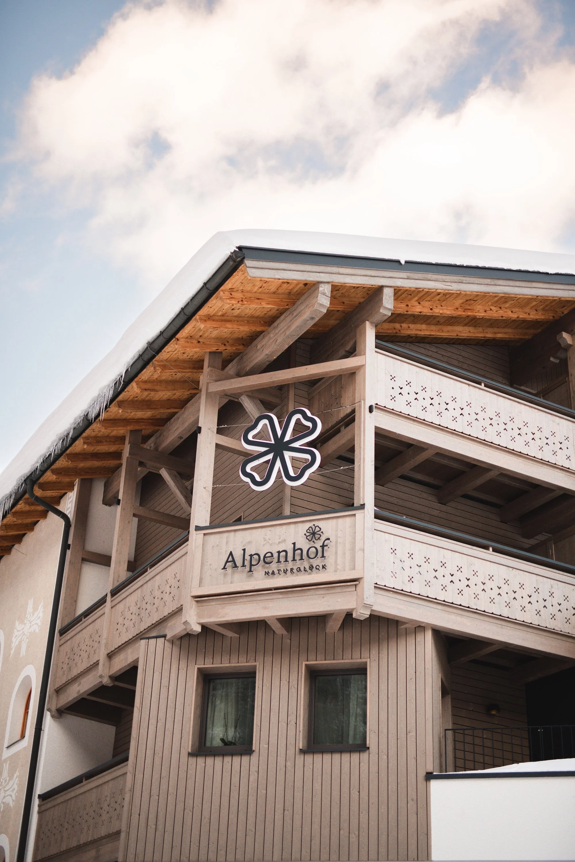 Mehrstöckiges Holzgebäude mit Balkon und Schneelast auf dem Dach, mit einem Laden namens 'Alpenhof NATURALPIS'.