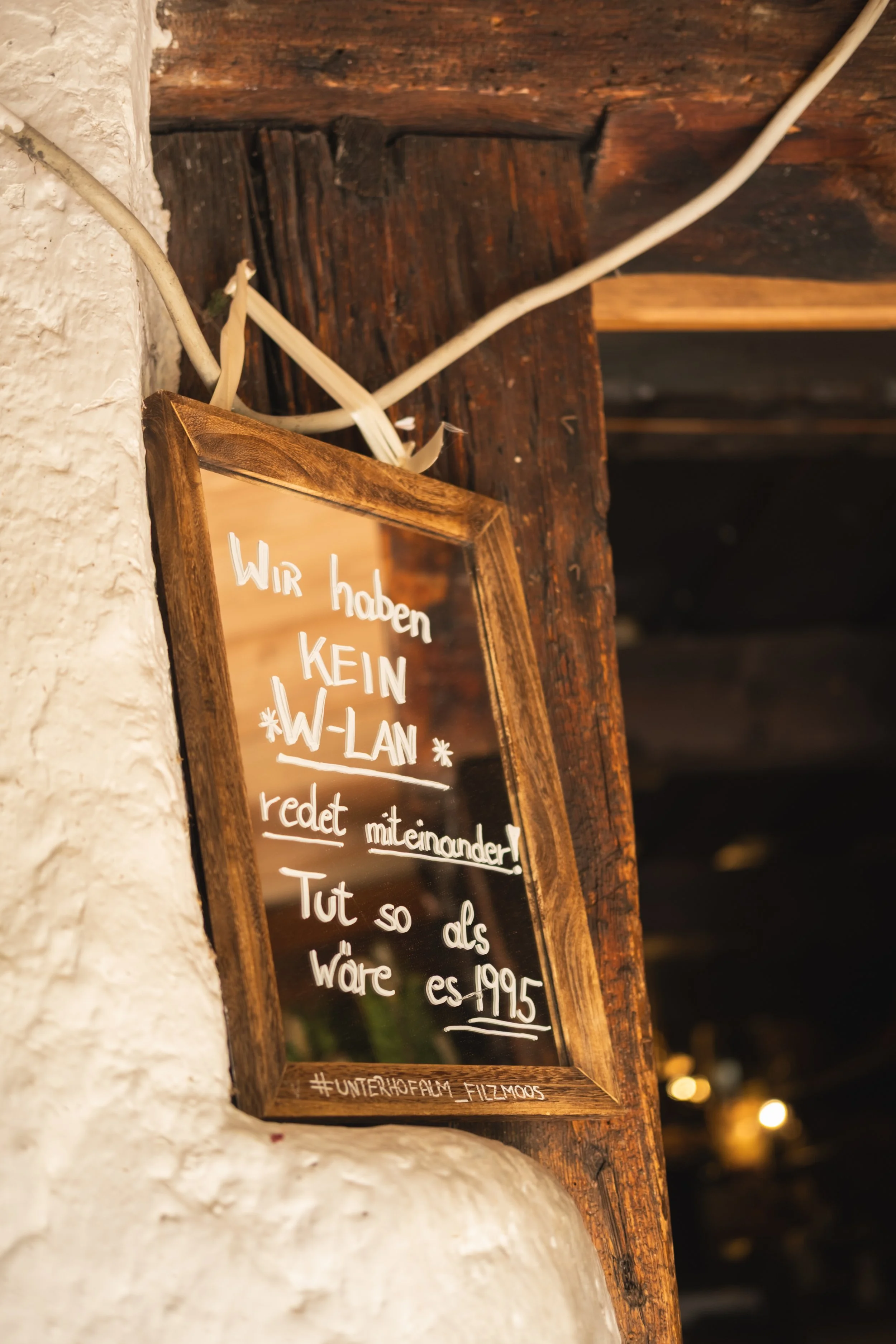 Ein Holzrahmen mit einem schwarzen Schreibbrett, auf dem in weißer Schrift geschrieben steht: "Wir haben KEIN W-LAN" und eine weitere Nachricht. Der Rahmen hängt an einer weißen Wand mit sichtbaren Noppen. Im Hintergrund sind unscharfe Lichter sichtb