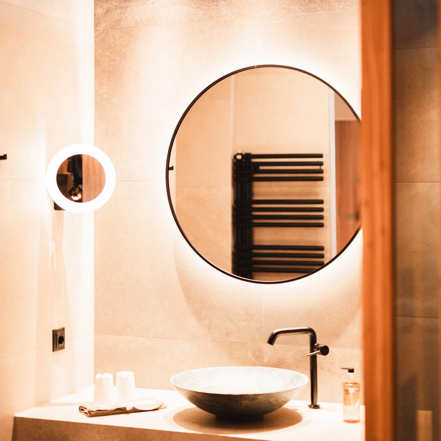 Modernes Badezimmer mit runder Spiegels, Waschbecken aus Sandstein, schwarze Wasserhähne, Handtuchhalter, Beleuchtung und Dekorationsartikeln.