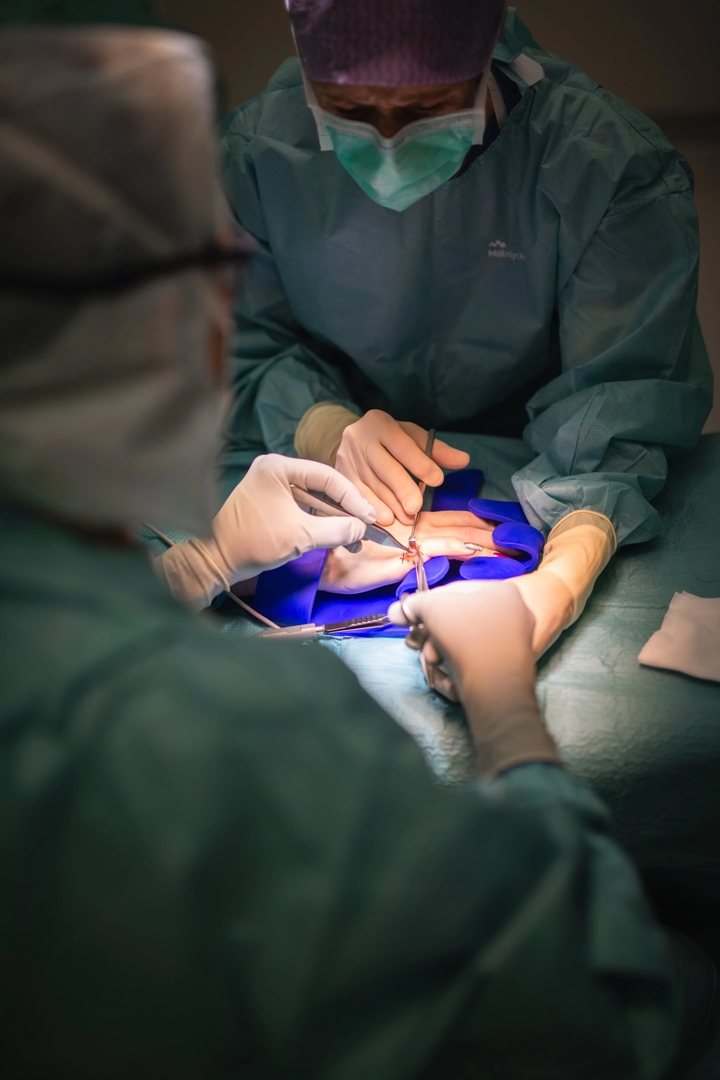 Chirurg bei einer Operation am Hand. Der Chirurg trägt eine grüne OP-Kleidung, Maske und Handschuhe und arbeitet mit einem Skalpell an dem Handgelenk des Patienten.