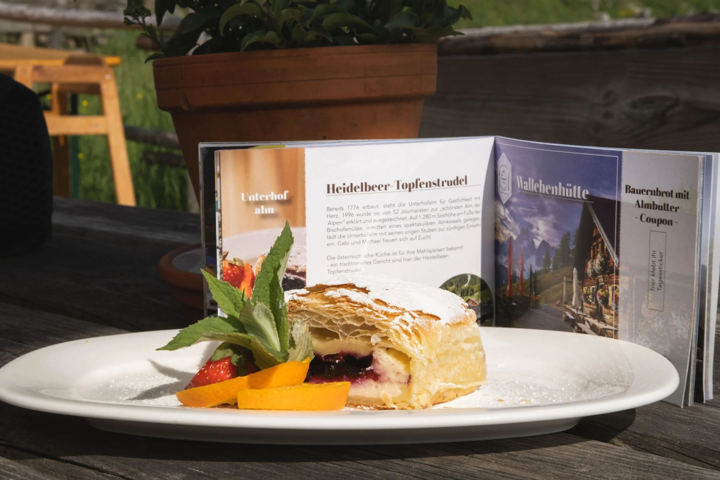 Ein Teller mit einem Stück Heidelbeer-Topfenstrudel, Früchten und frischer Minze. Im Hintergrund eine Speisekarte mit Beschreibung des Strudels, auf einer Holzbank im Freien.