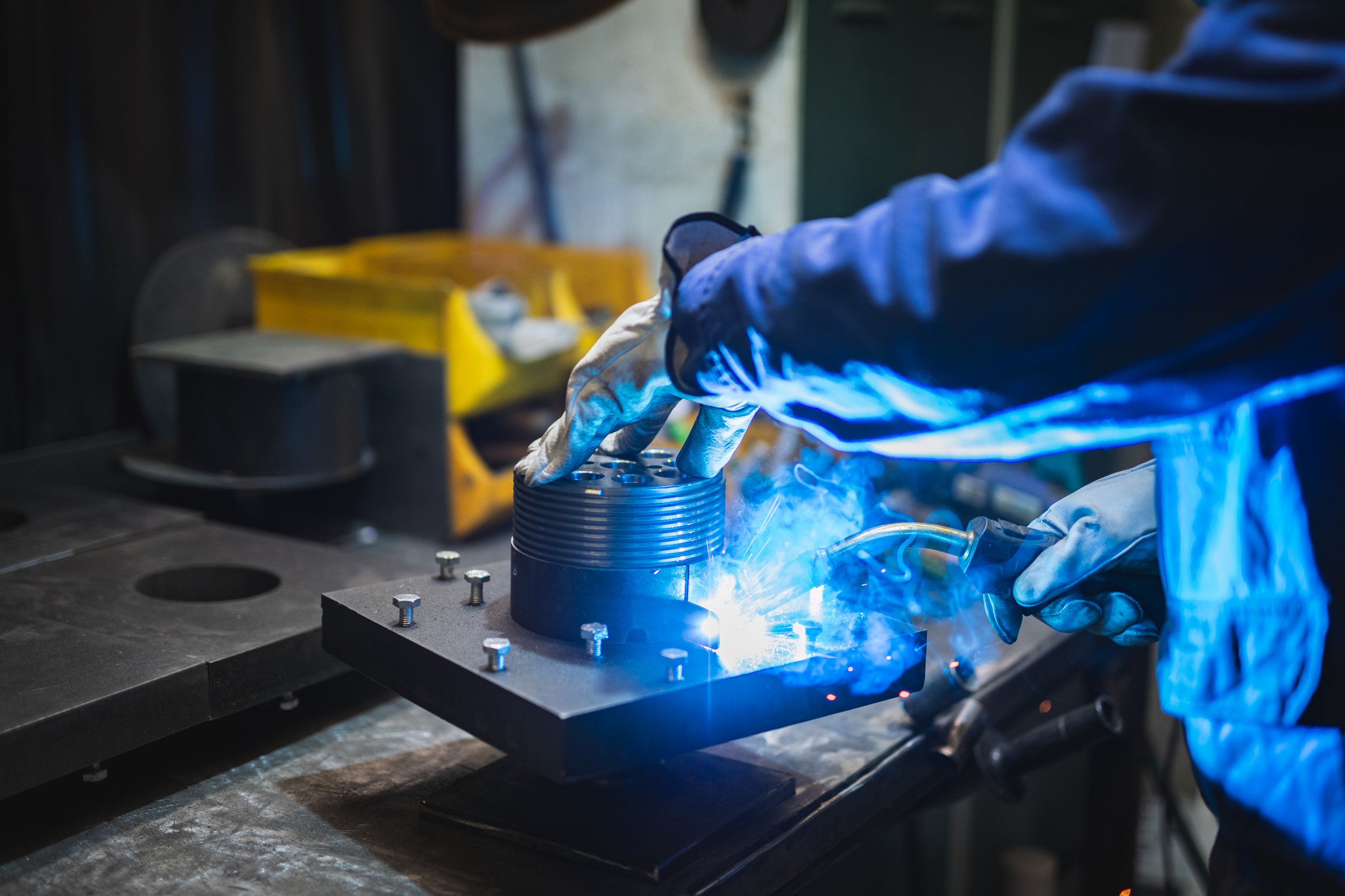 Ein Arbeiter trägt eine Schutzbrille und Handschuhe und schweißt ein Metallstück auf einer Werkbank. Es ist eine industrialle Werkstatt mit Werkzeugen und Schrauben auf der Oberfläche.