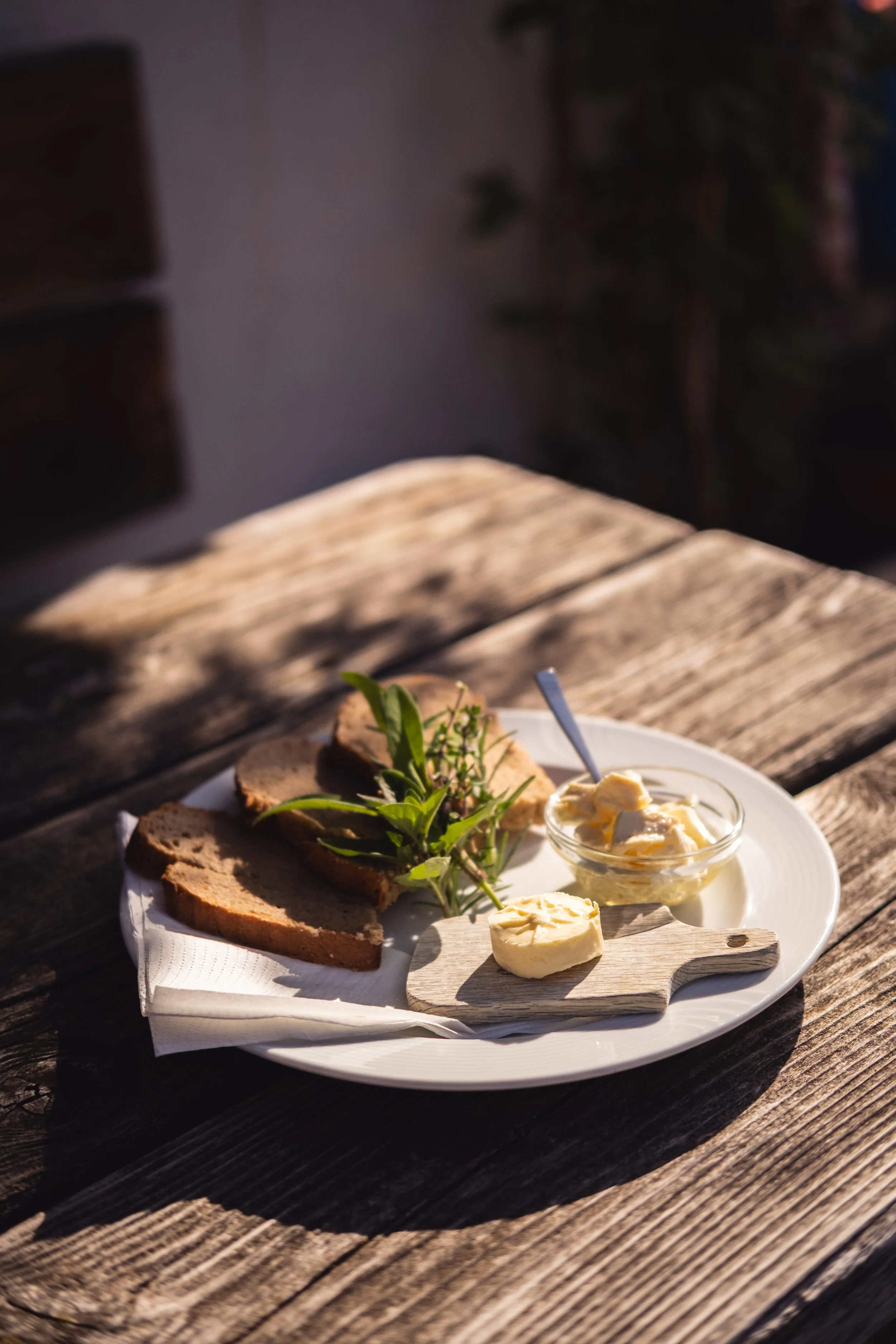 Brot, Käse, Butter, Salat auf einem weißen Teller auf einer Holztischfläche, im Hintergrund unscharf.