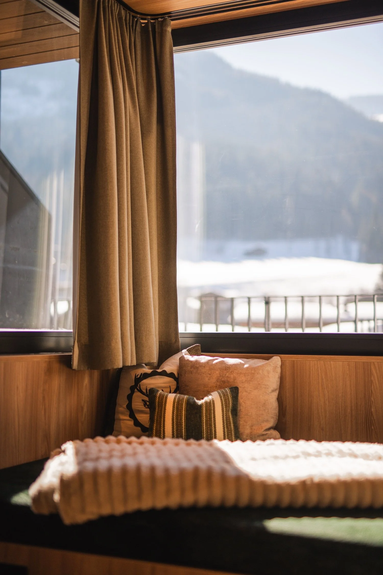 Blick auf eine Fensterbank mit Kissen und Decke, vor einem großen Fenster mit Blick auf verschneite Berge und eine Terrasse im Winter.