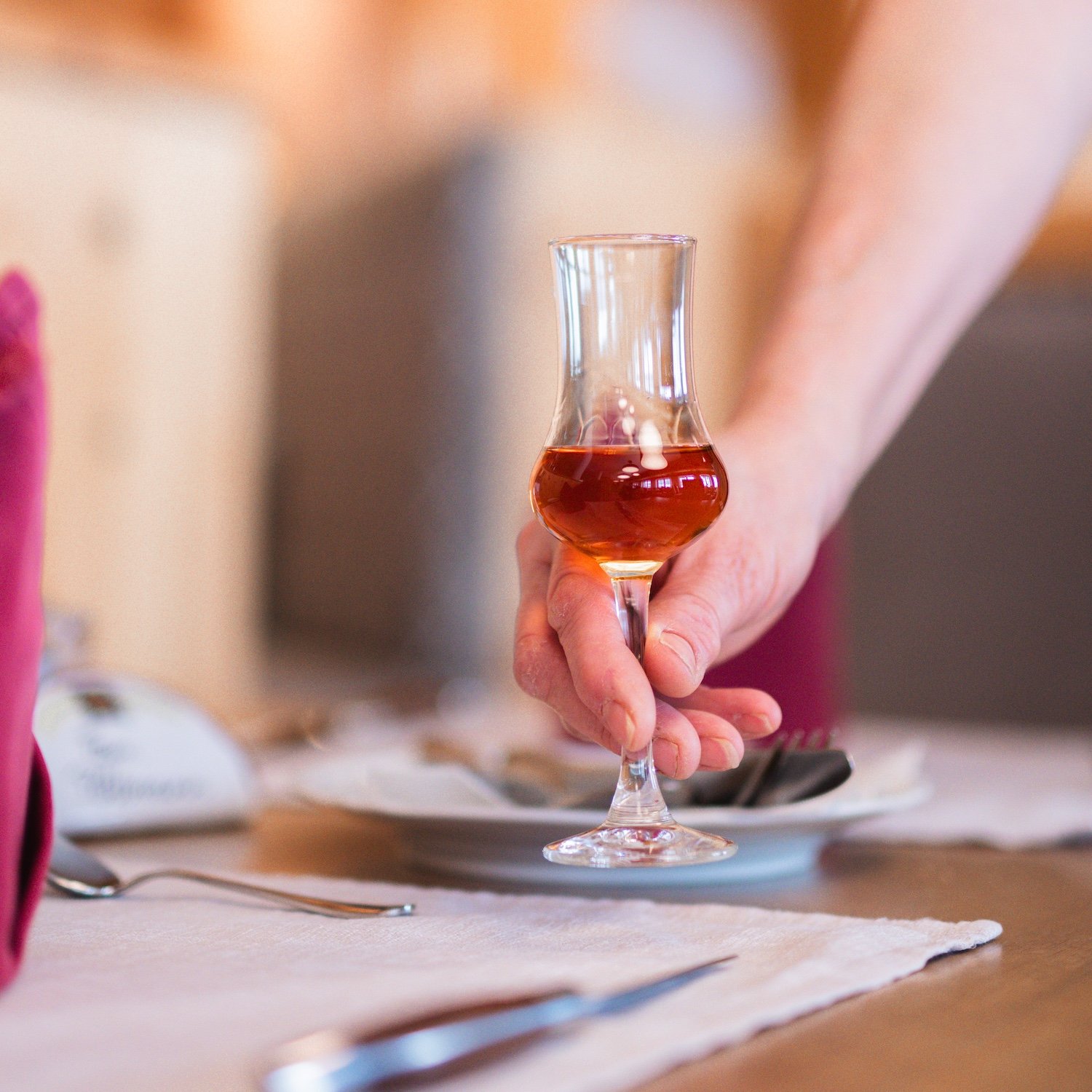 Person hält ein Cocktailglas mit braunem Getränk, wahrscheinlich einen Martini, in einem Restaurant oder zu Hause.