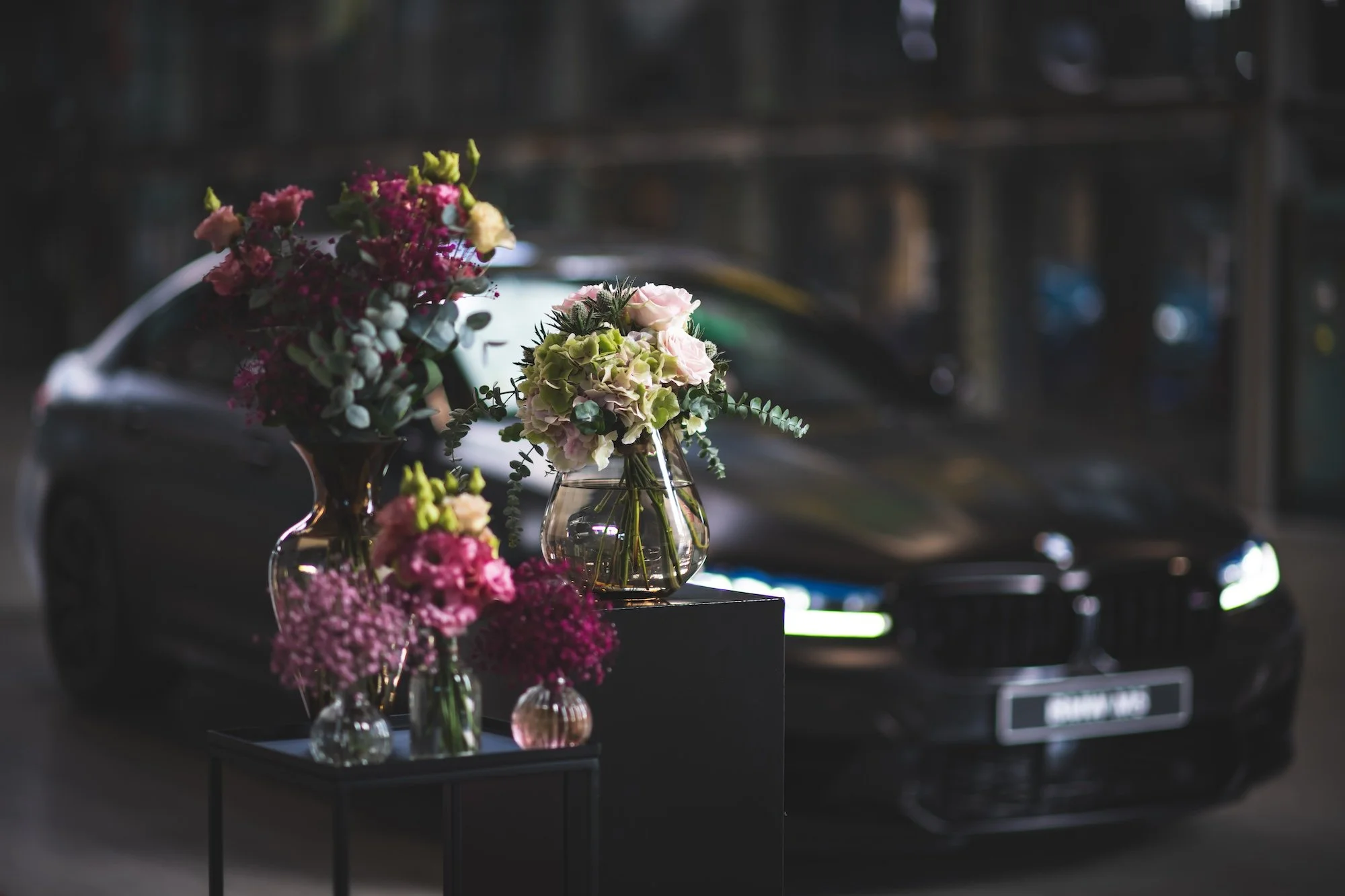 Blumenarrangements in Vasen auf schwarzen Tischen in einer dunklen Umgebung, wahrscheinlich in einem Ausstellungsraum oder auf einer Veranstaltung.