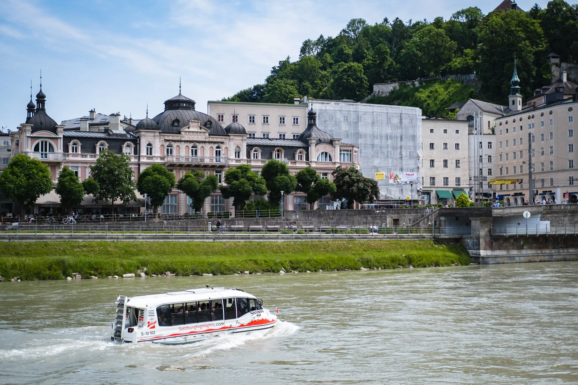 Auf einem Fluss fährt eine weiße Touristenfähre mit roten Schriftzügen, im Hintergrund elefantenförmige historische Gebäude und eine bewaldete Anhöhe. Verschiedene Bäume säumen den Flussufer.