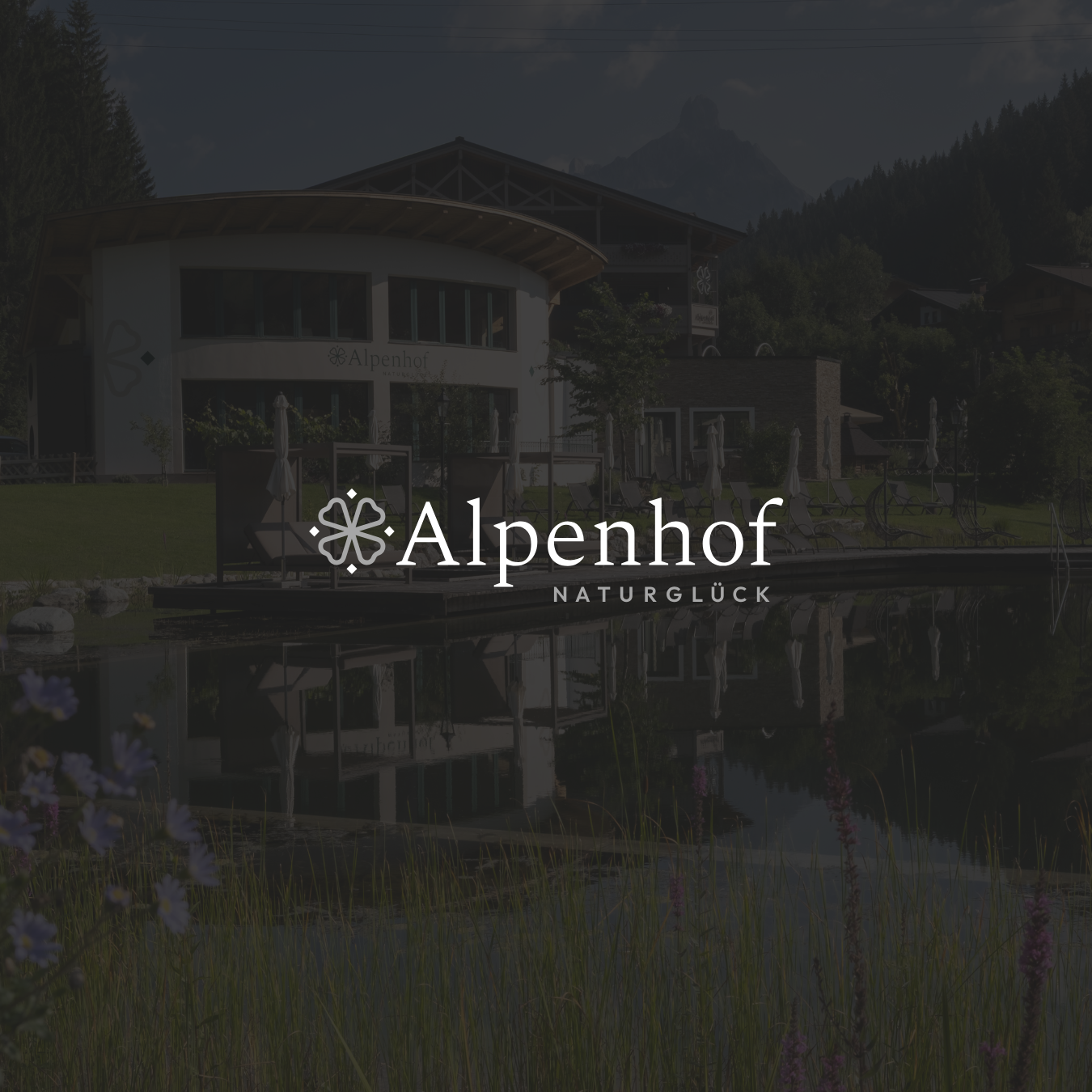 Ein modernes Gebäude mit einem Wasserteich im Vordergrund, umgeben von Bänken und Liegestühlen, mit Bergen im Hintergrund, die den Alpenhof Naturglück repräsentieren.