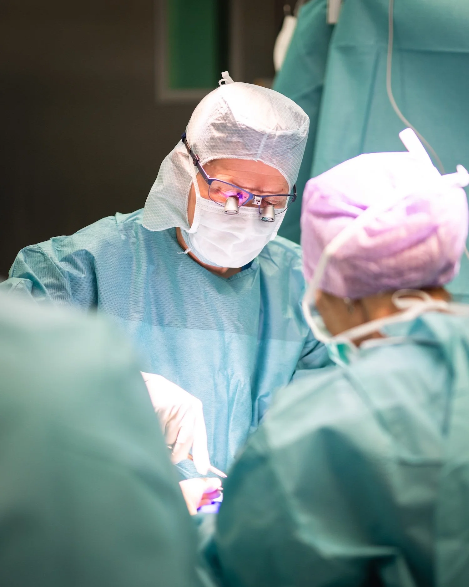 Ärzte und Pflegepersonal bei einer Operation im Operationssaal, überwiegend in grüner OP-Kleidung. Die Hauptperson trägt eine Schutzmütze, eine OP-Maske und eine Lupenbrille.