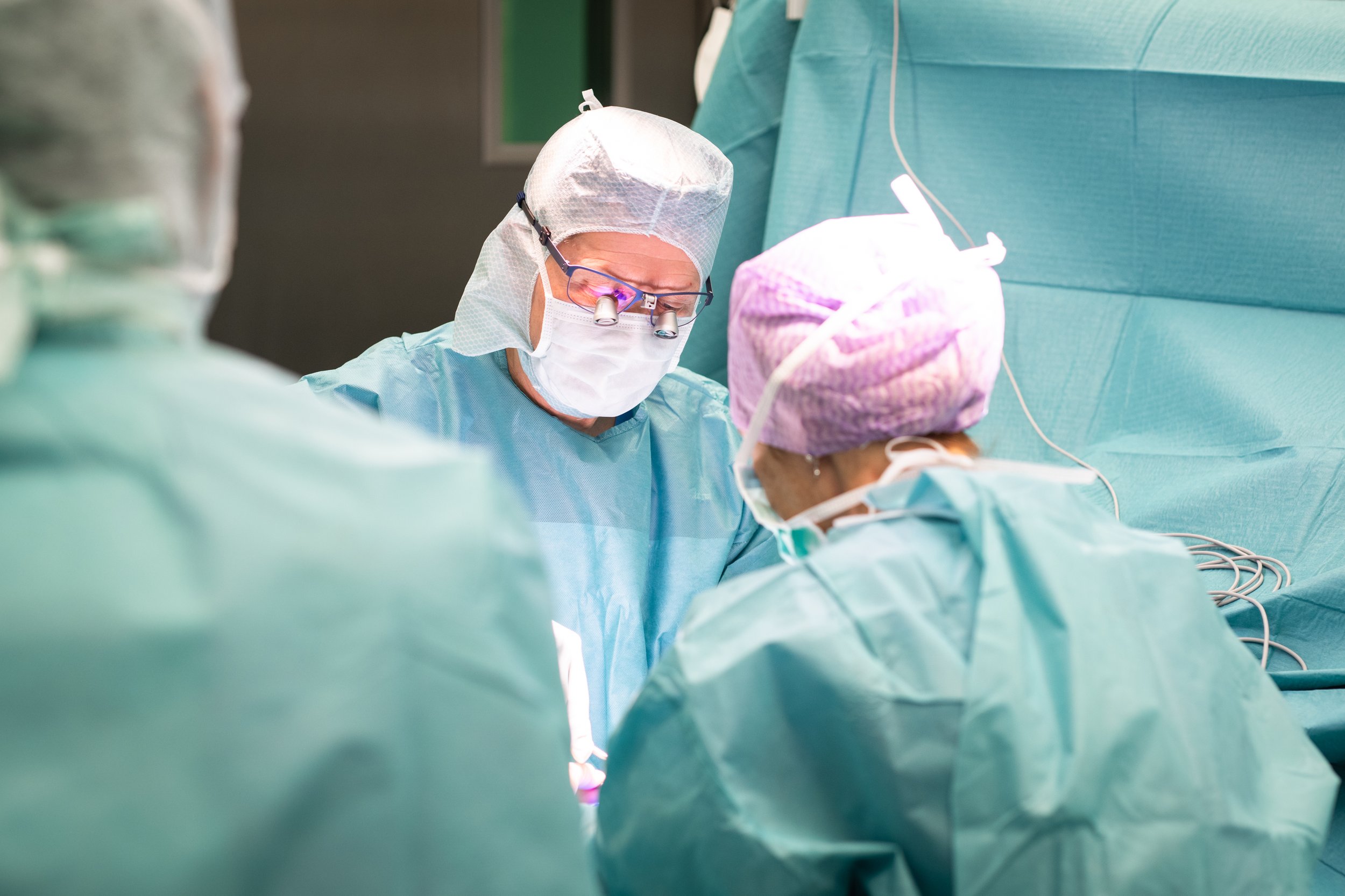 Chirurgen während einer Operation in einem Operationssaal, gekleidet in blaue OP-Kleidung, Maske, Schutzbrille und Haarschutz. Sie arbeiten fokussiert an einem Eingriff.