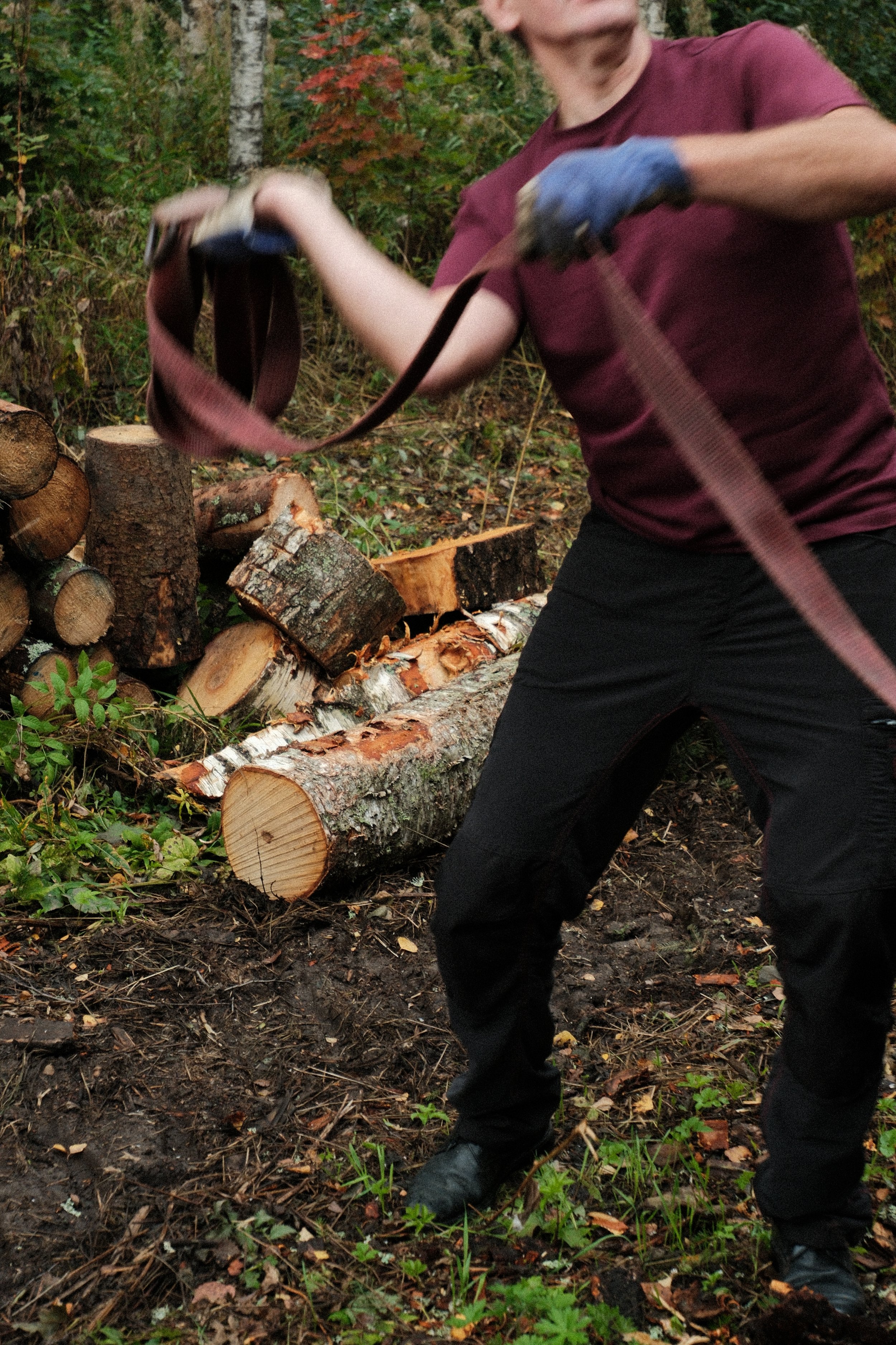 A person securing a strap on the cargo in a wooded area, wearing a maroon shirt and black pants, with logs and chopped wood visible in the background.