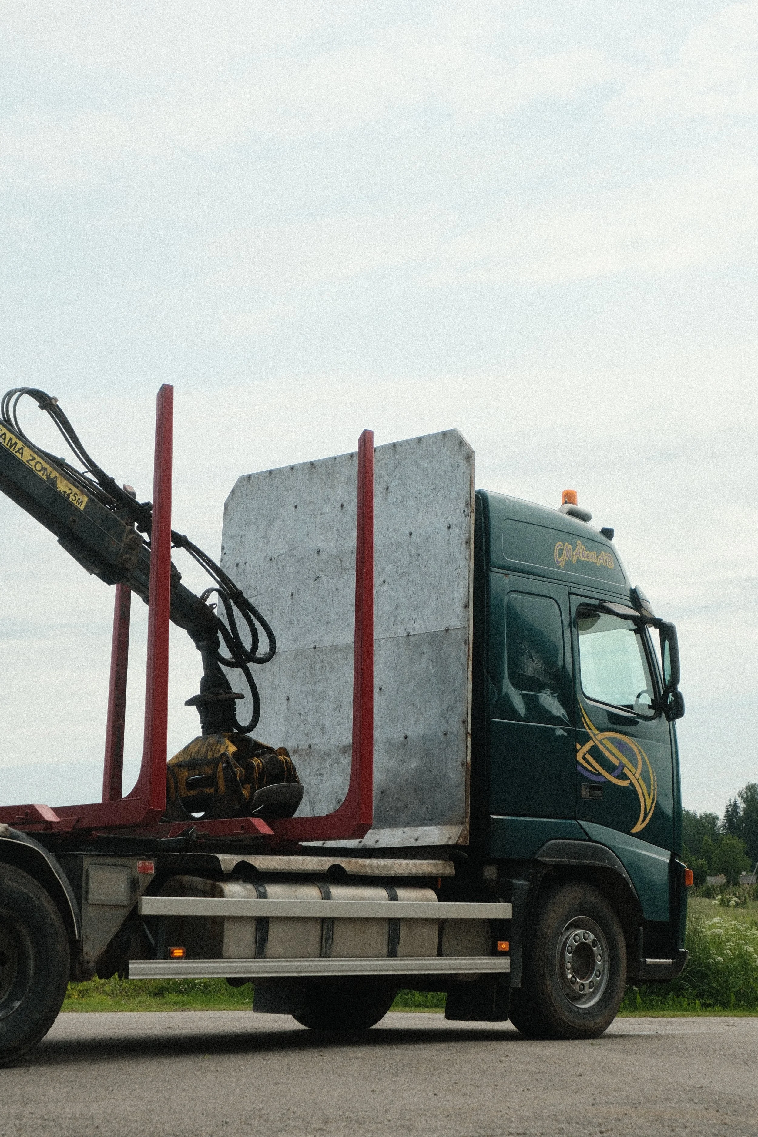 Side view of a semi-truck with a dark green cab, a large white panel, red vertical supports, and a hydraulic arm attached, parked on a road with trees and cloudy sky in the background.