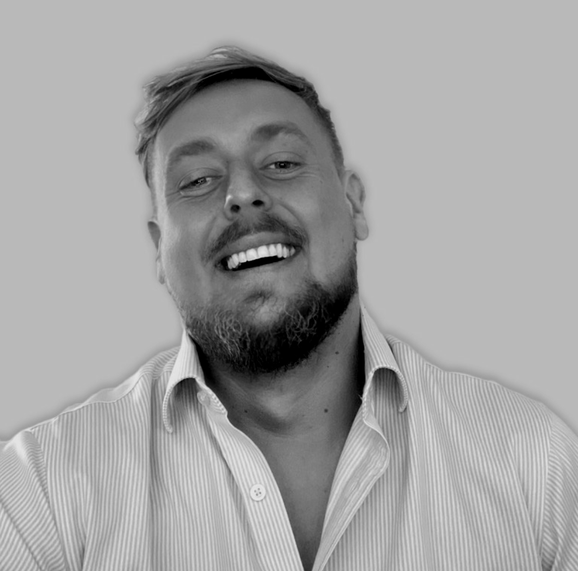 Black and white portrait of a smiling man with short hair, beard, and mustache, wearing a collared shirt with vertical stripes, against a plain background.