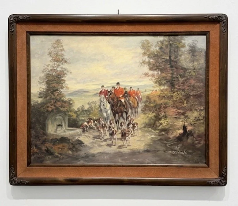 A framed painting depicting a group of fox hunters riding horses and accompanied by a pack of foxhounds through a forested landscape, with trees and a stone structure in the background.