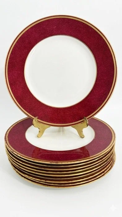 A set of decorative plates with gold rims, stacked, with a solitary plate propped open showing a red and gold border design.