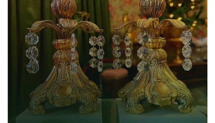 Two ornate vintage brass candelabras with crystal hanging decorations on a reflective surface, with a blurred decorated Christmas tree in the background.