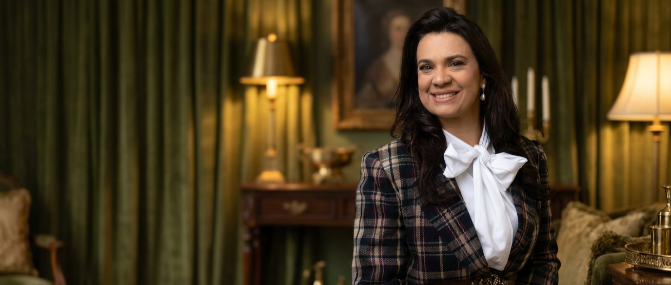 A woman with dark hair smiling in a vintage-style room with green curtains, lamps, and framed artwork.