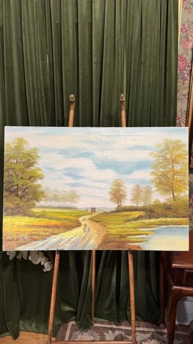 Landscape painting of a dirt path through green fields with trees and a cloudy sky, on an easel in front of a green curtain.