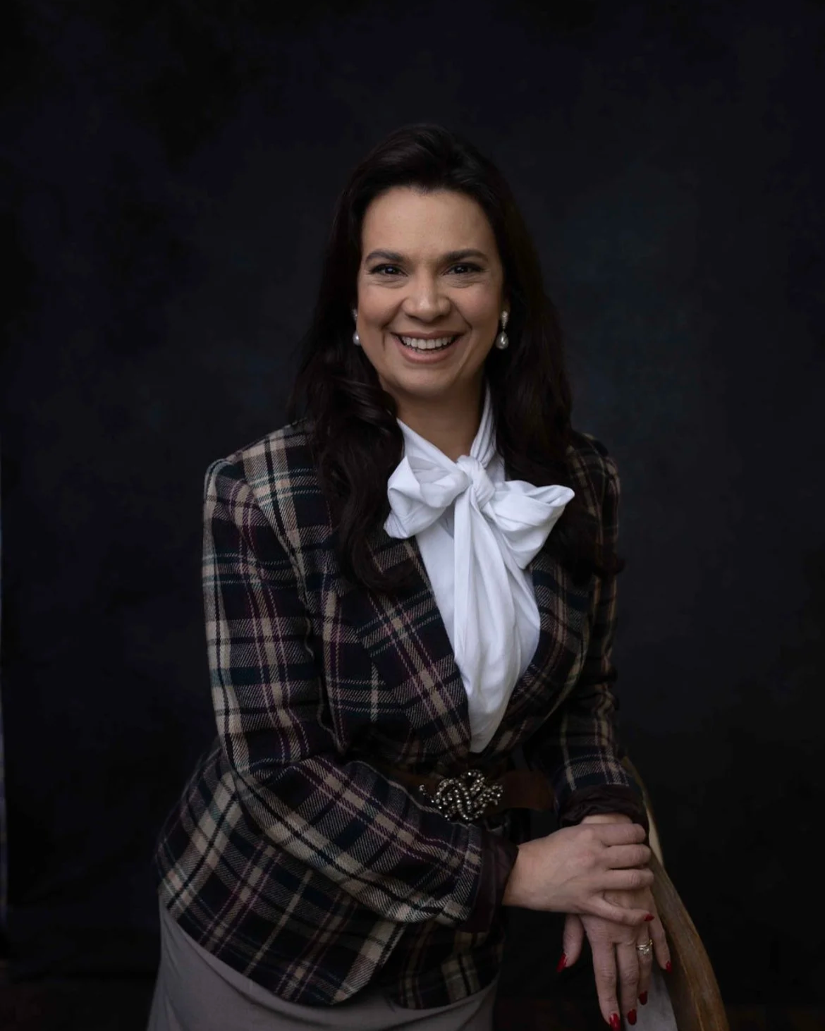 A woman with dark brown hair smiling, wearing pearl earrings, a white blouse with bow tie, and a brown plaid blazer, sitting against a dark background.
