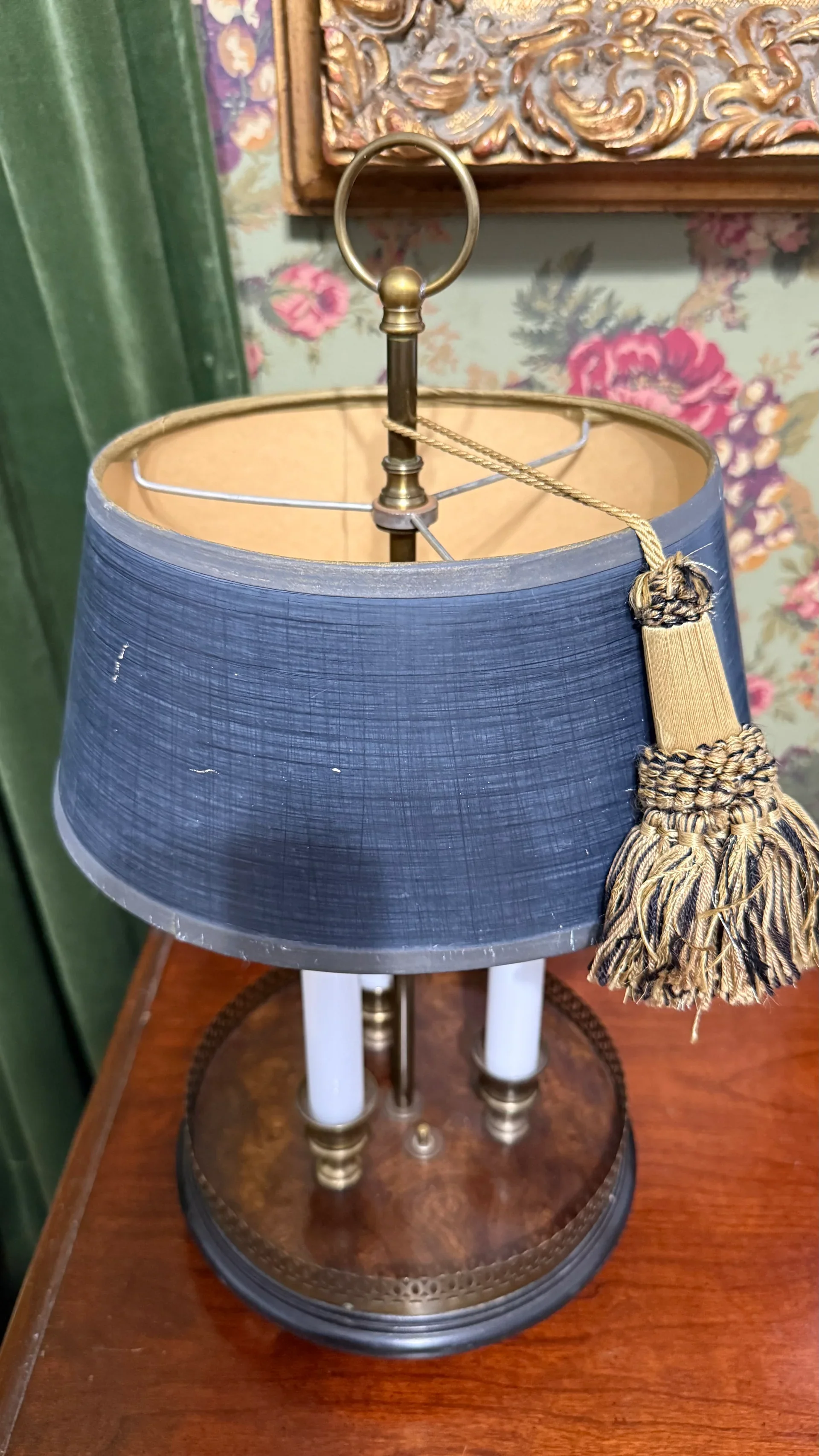 A vintage table lamp with a blue fabric shade, gold accents, and a decorative tassel hanging from the top, positioned on a wooden surface.