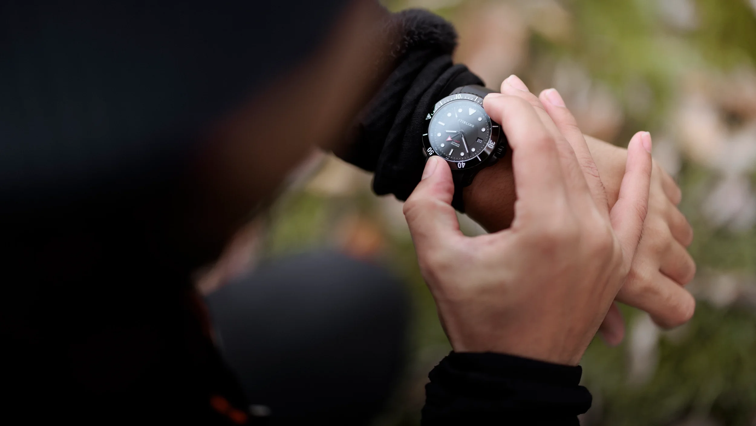 A person adjusting a black wristwatch on their left wrist outdoors, with blurred green and brown background.