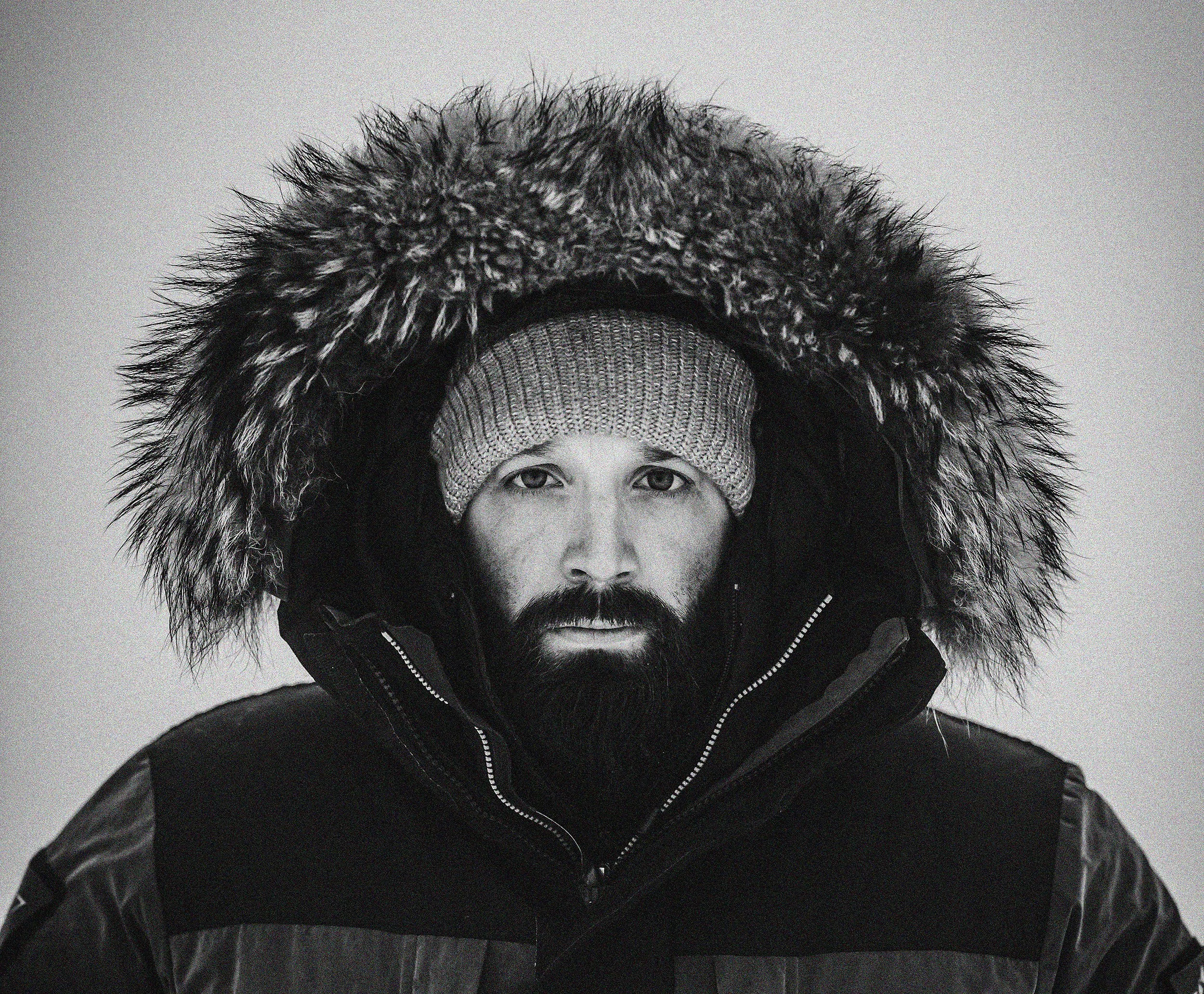 Close-up of a person wearing a warm winter jacket with a large fur-lined hood, a knit beanie, and a scarf, with half of their face visible and looking into the camera.