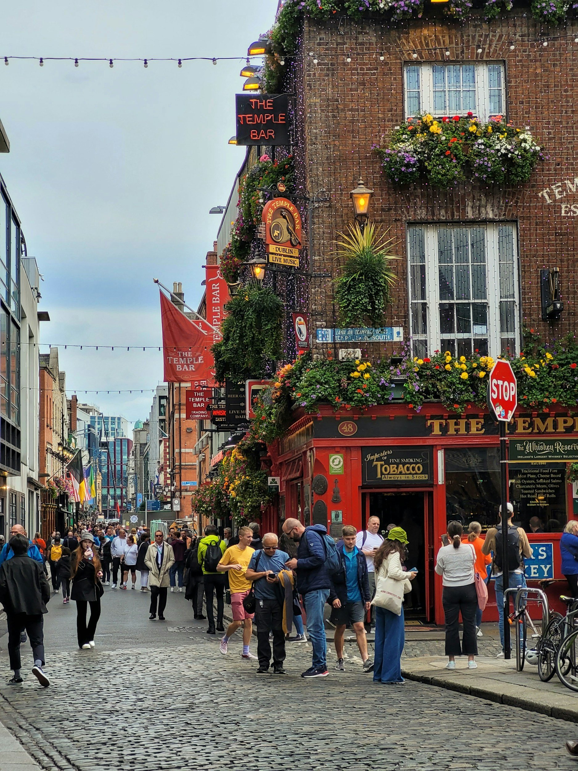 Crowds of people walking along a busy city street with shops and colorful flowers, signage for The Temple Bar, and flags visible.