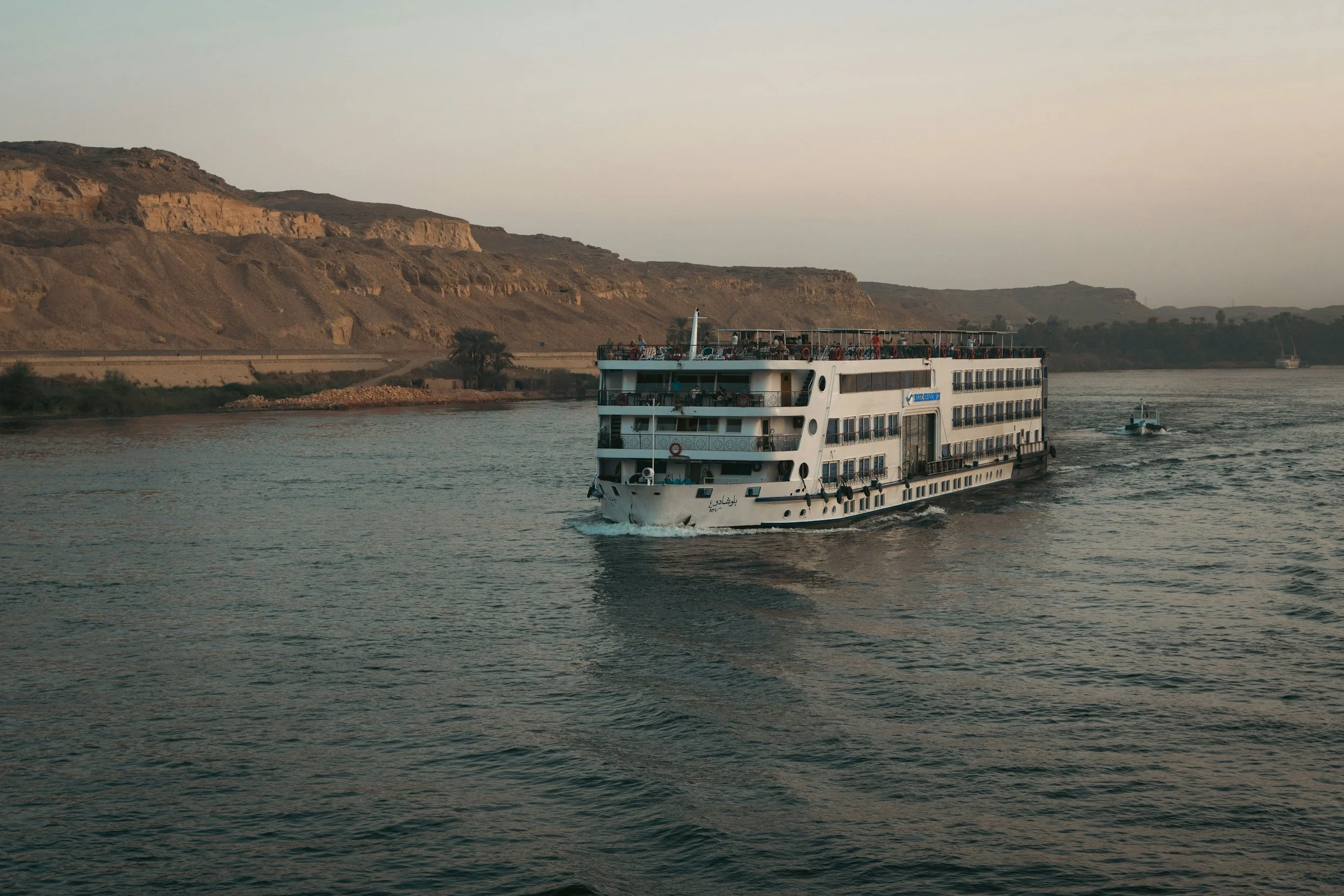A white multi-deck cruise ship sailing on calm river waters near a brown, rocky hillside with sparse vegetation.