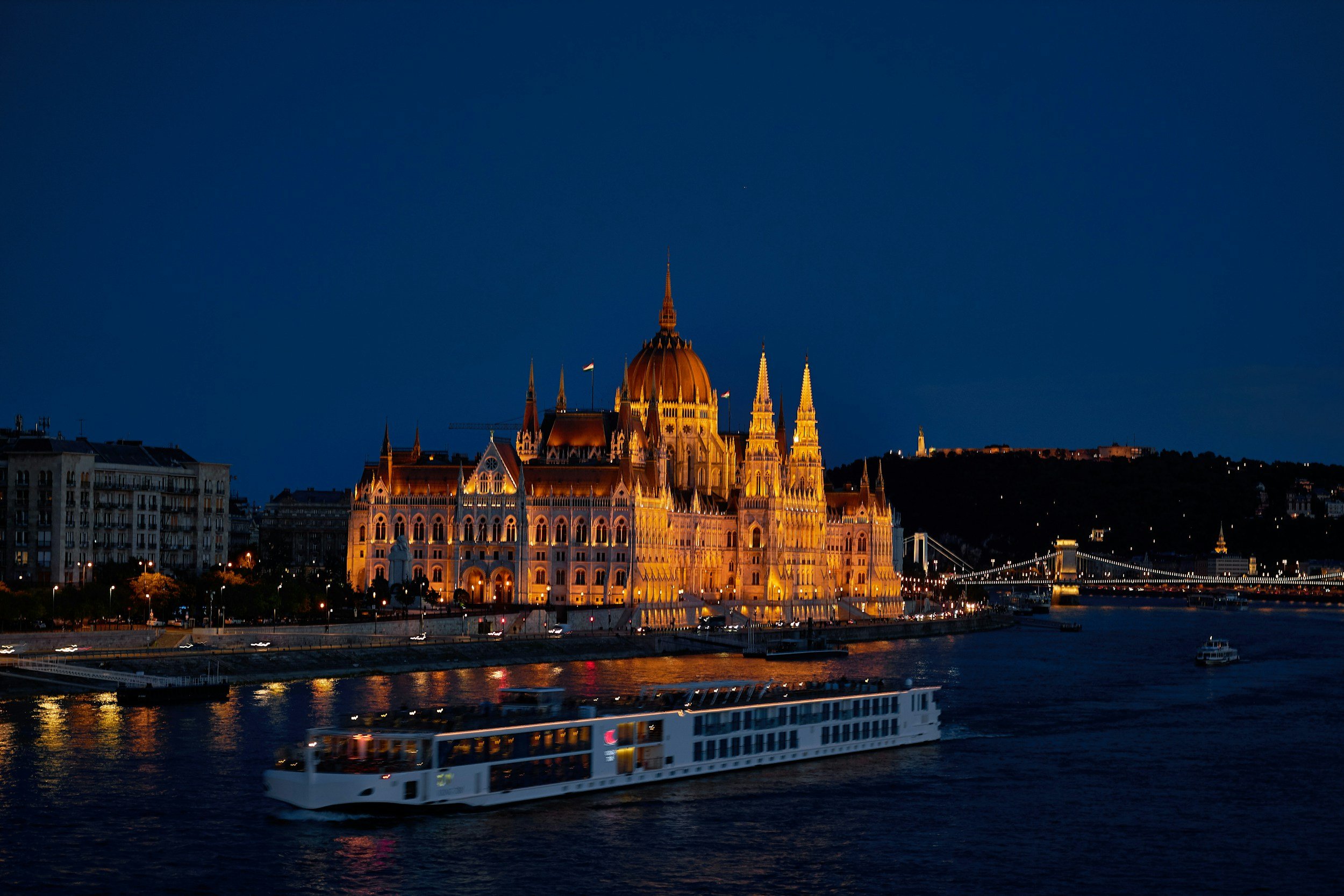 Night view of the Hungarian Parliament Building in Budapest illuminated, with the Danube River in the foreground and a boat passing by, and the Chain Bridge lit in the background.