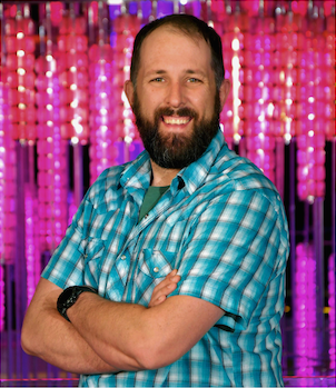 A man with a beard wearing a blue plaid shirt smiling with arms crossed, against a pink and purple glittery background.
