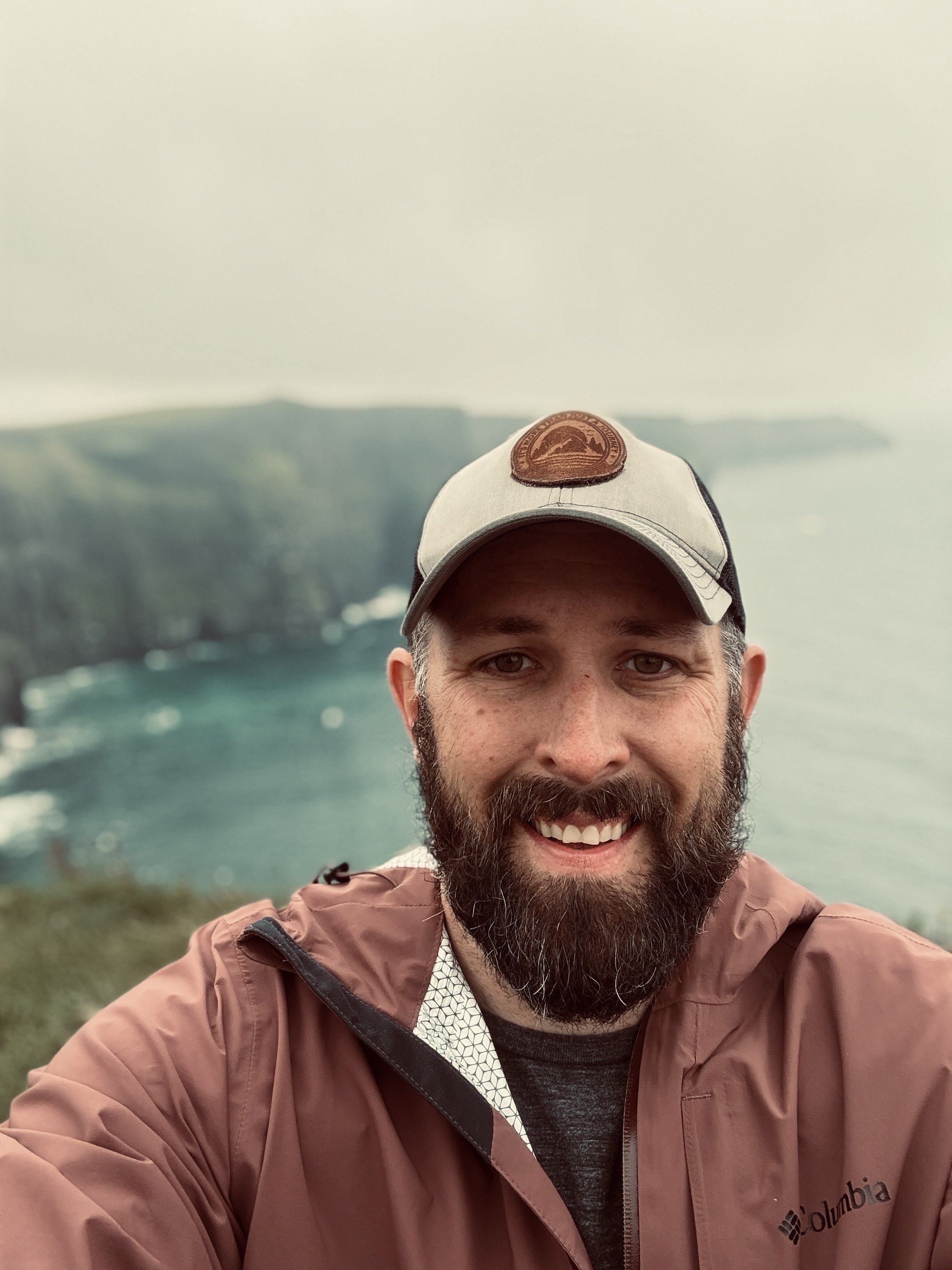 A smiling man with a beard and mustache wearing a Columbia hat and jacket, taking a selfie with a scenic mountain and water background.