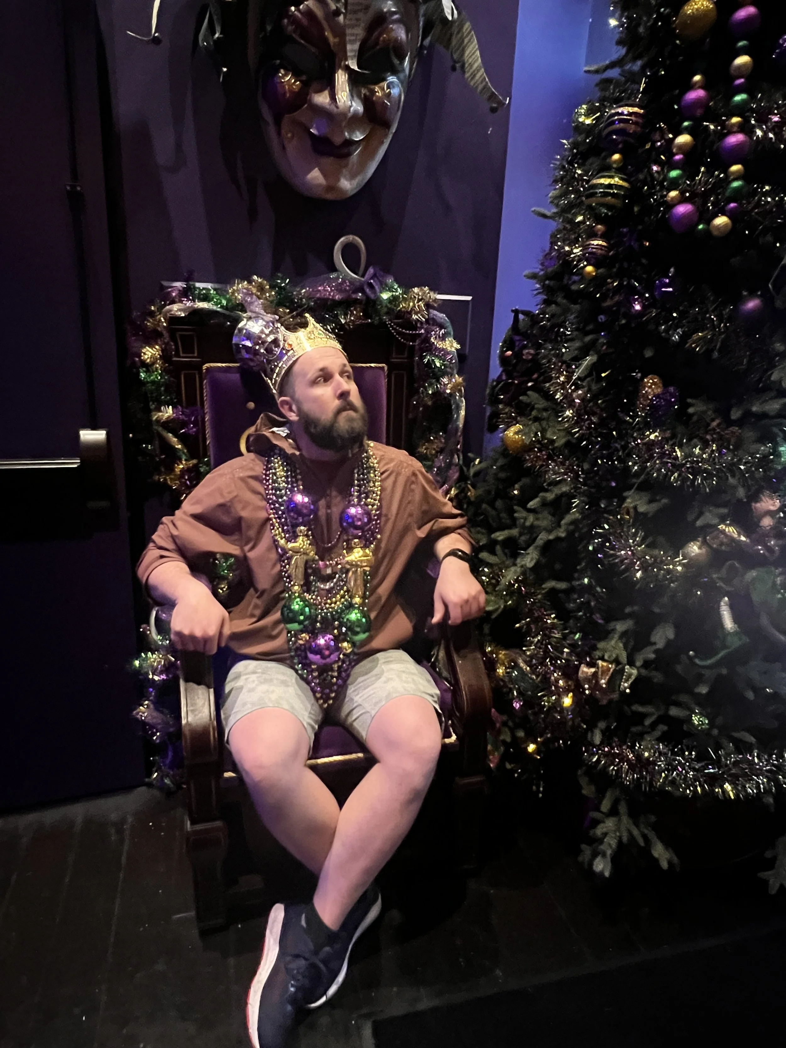 A man with a beard sitting in a throne surrounded by Christmas decorations, wearing a crown and multiple bead necklaces, with a decorated Christmas tree beside him.