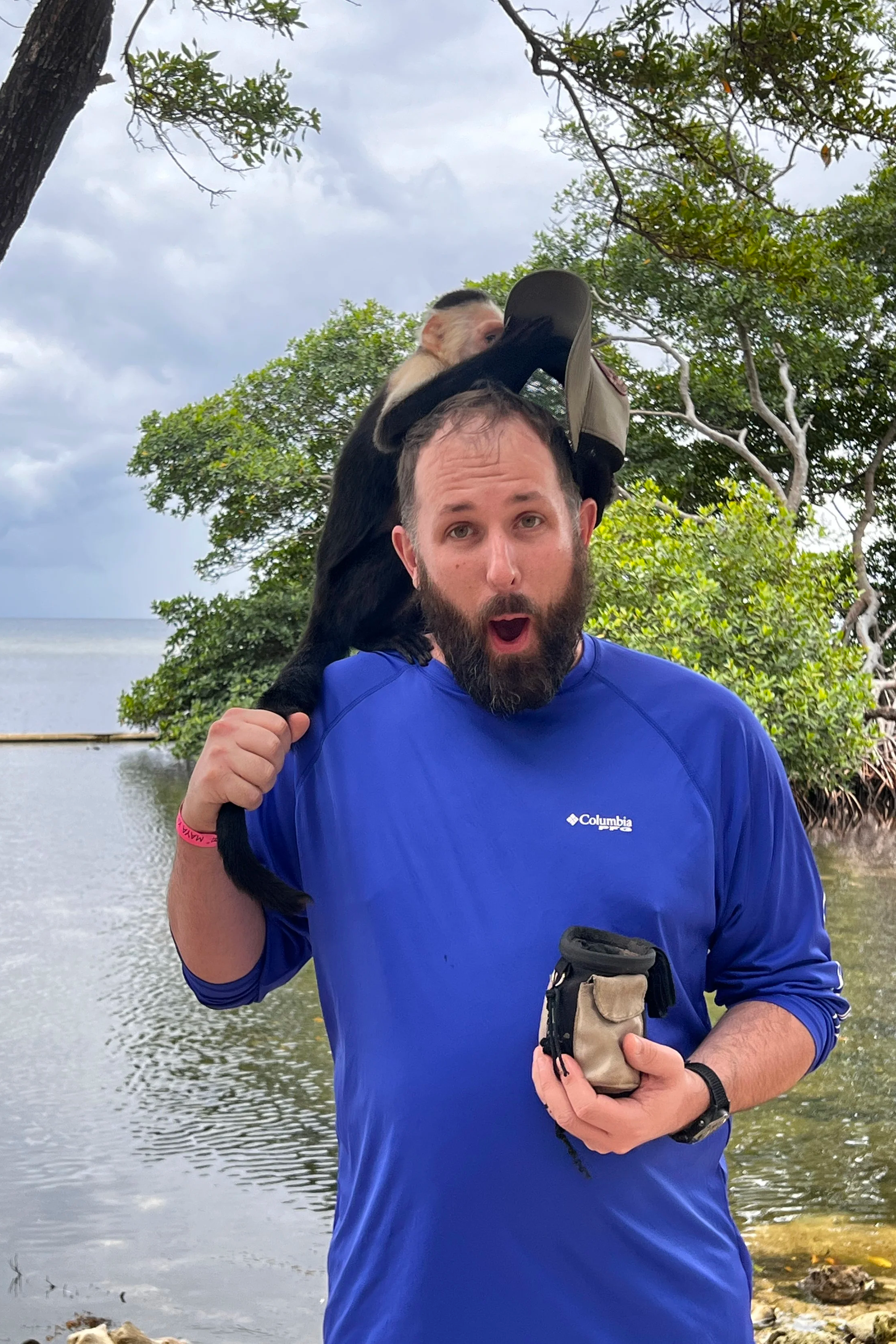 A man with a beard and blue Columbia shirt holding a small glove in front of him, with a monkey on his shoulder in a tropical outdoor setting with trees and water in the background.