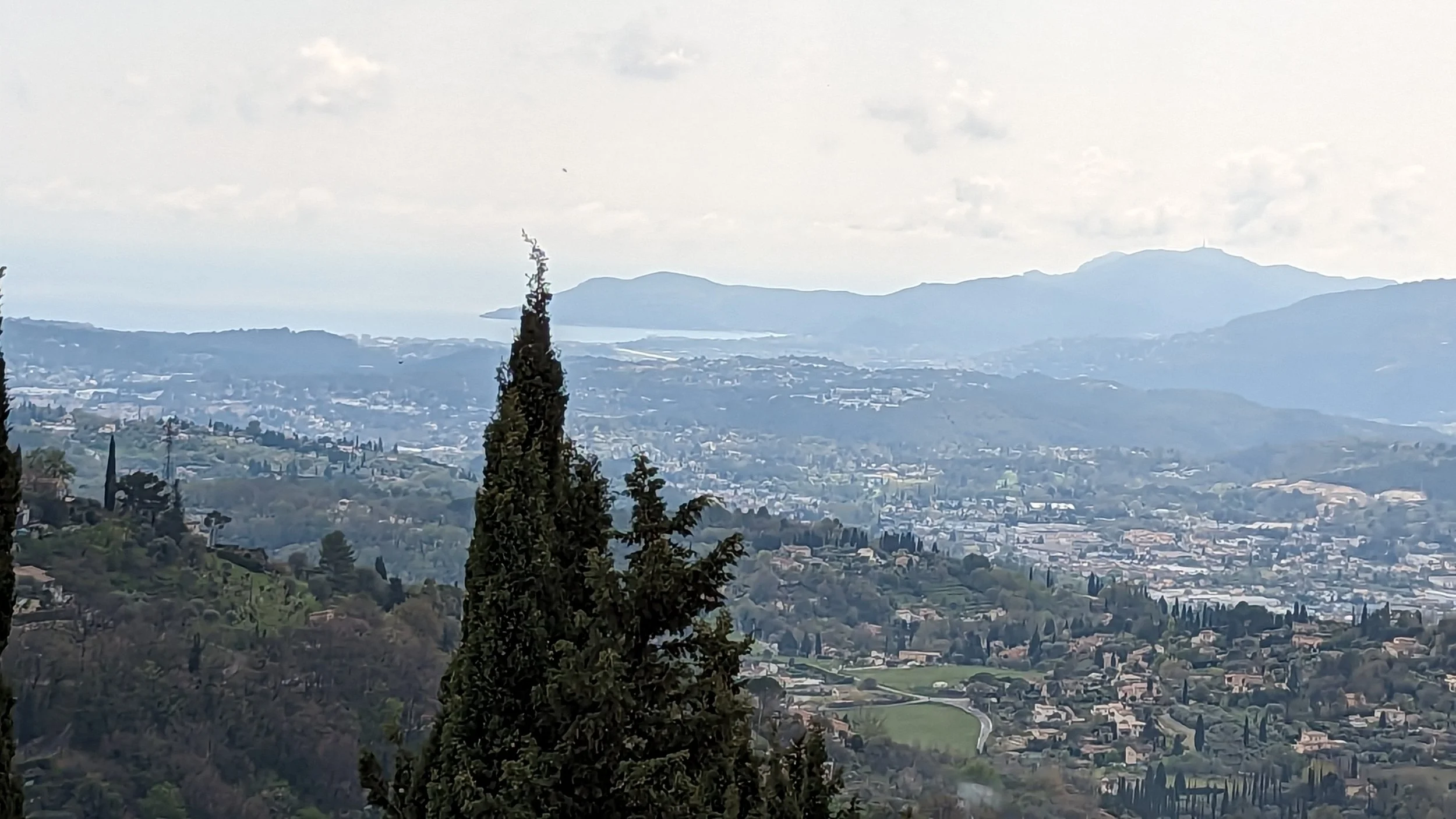 A scenic view of a hilly landscape with dense trees, a town with numerous houses, and mountain ranges in the background under a cloudy sky.