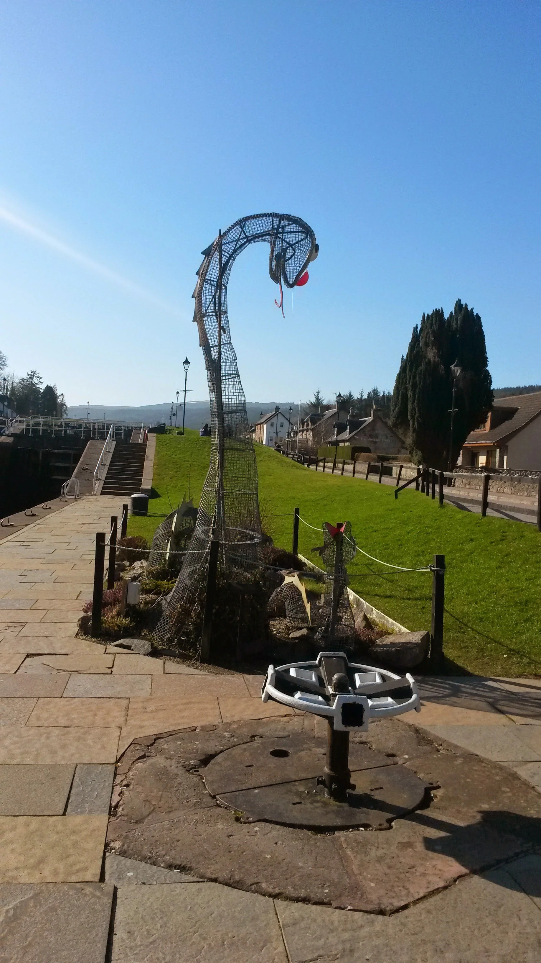 A decorative metal sculpture of a swan with a red beak and neck, situated in a grass-covered park area. There is a pathway and a bike station in the foreground, with houses, trees, and a clear blue sky in the background.