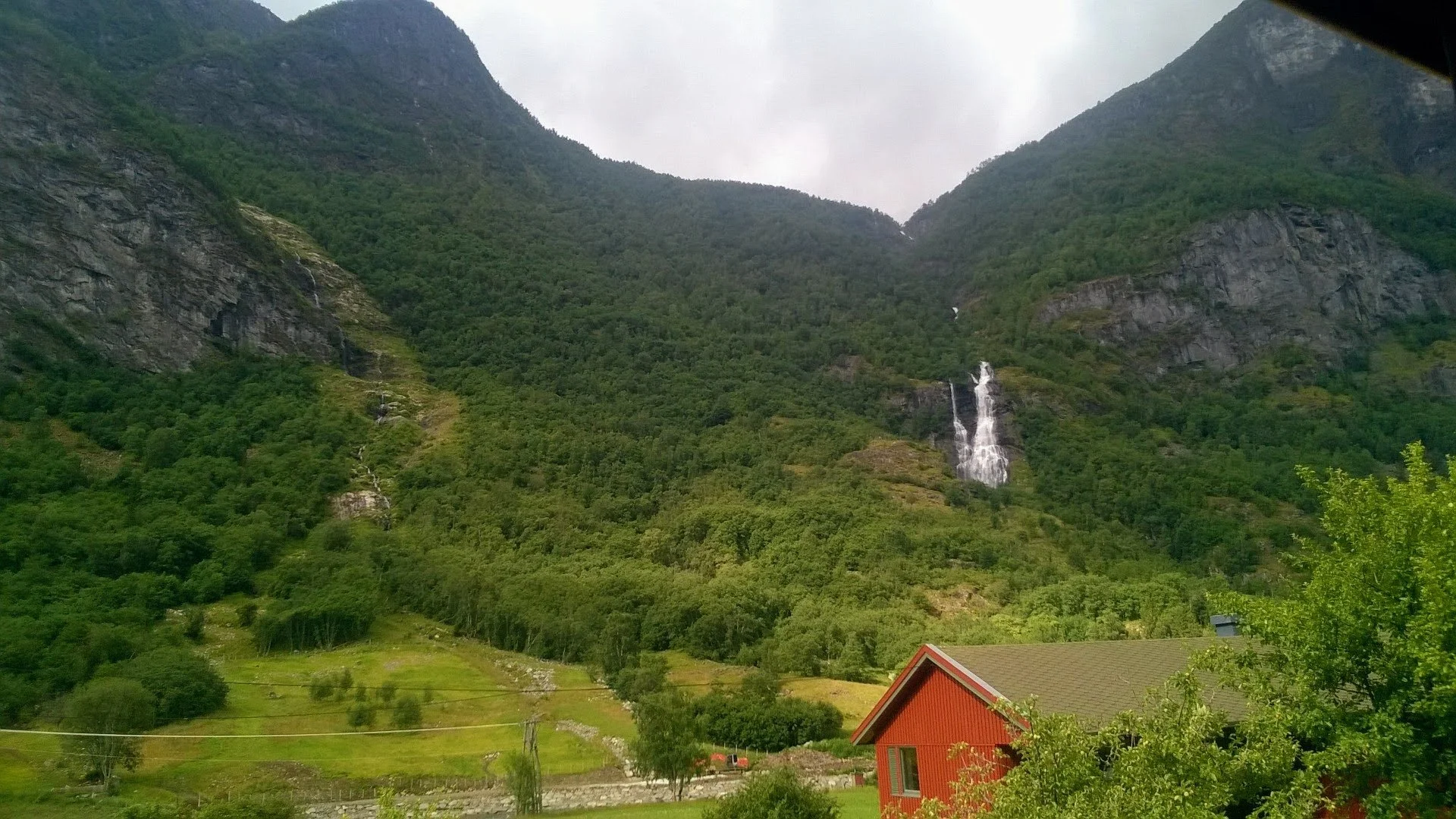 A scenic view of green mountains with a small waterfall cascading down one side. There is a red house with a gray roof in the foreground and some trees and telephone poles below the mountains.