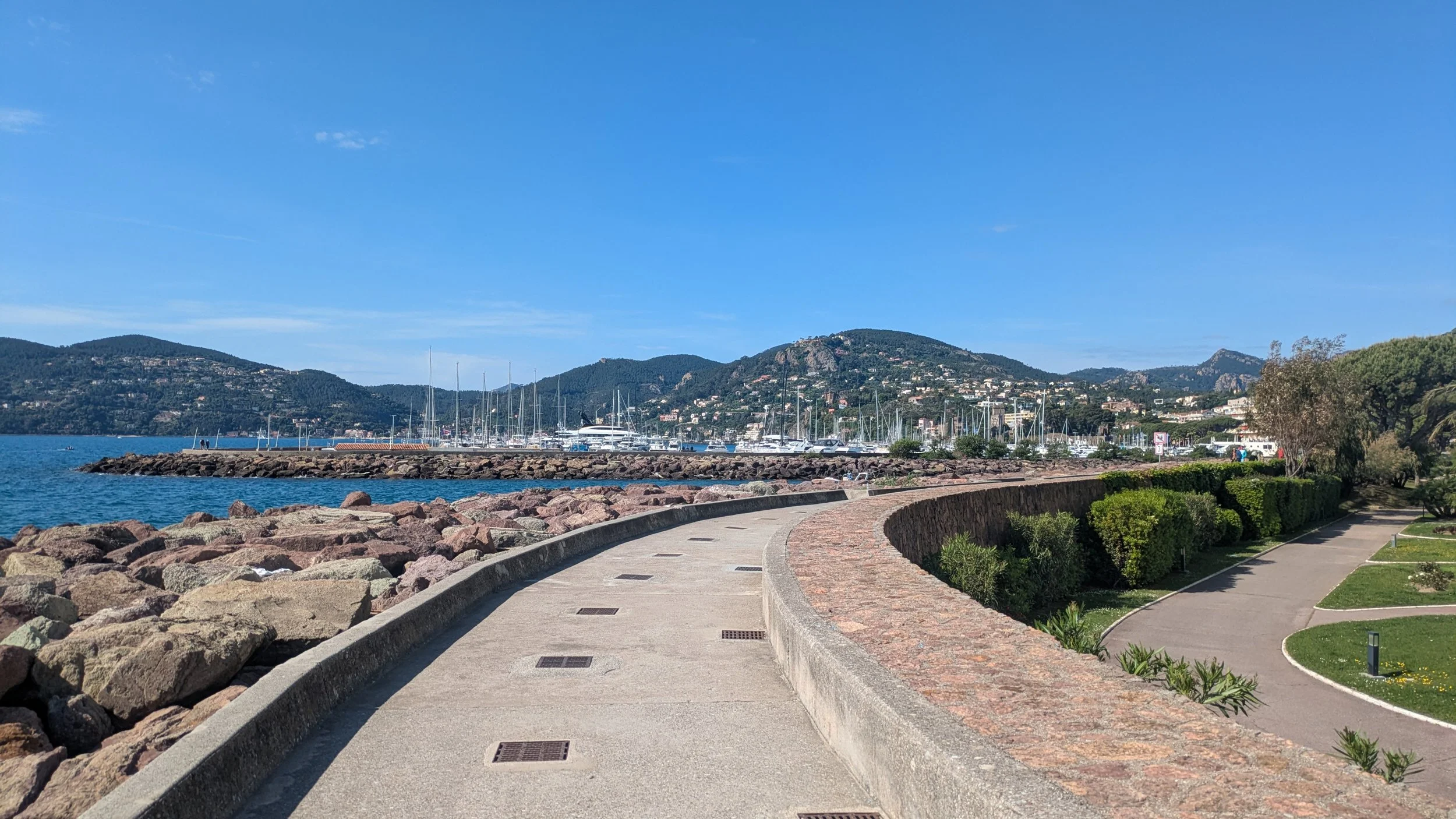 Scenic view of a marina with sailboats docked, a rocky breakwater, a curved concrete walkway, green bushes, trees, hillside with houses, and a blue sky.