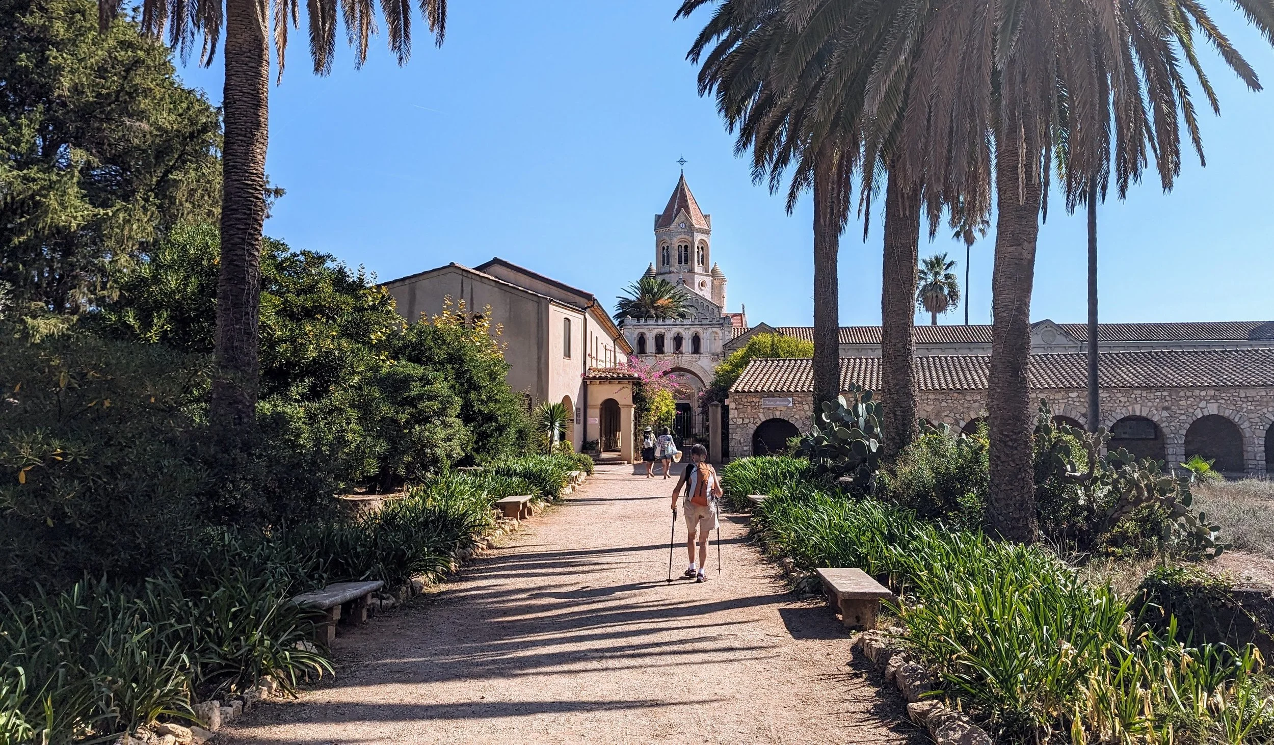 A narrow dirt path lined with green plants and bushes leads towards a church with a tall bell tower, surrounded by tall palm trees and other greenery, under a clear blue sky.