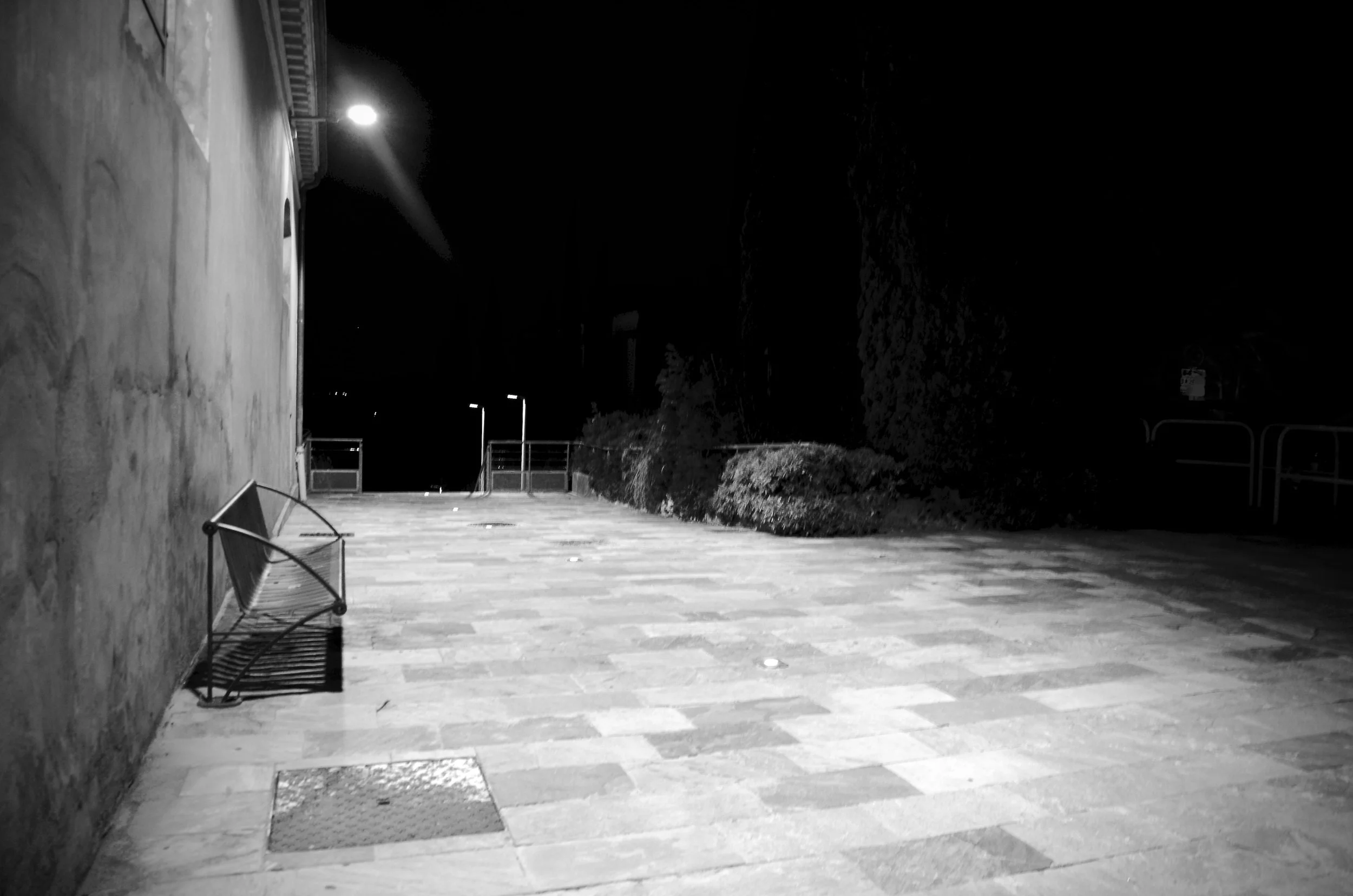Empty outdoor patio at night with two metal benches, a concrete wall, bushes, and trees in the background, illuminated by a single outdoor light.