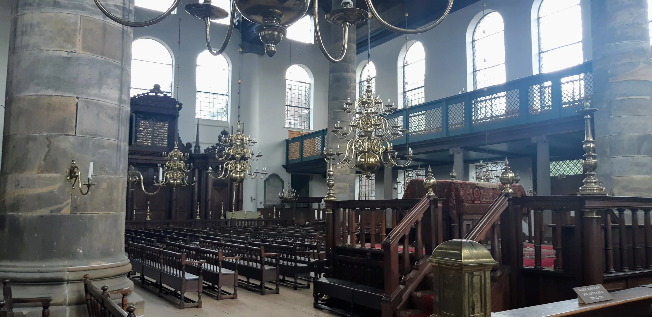 Interior of a church or historic building with large stone columns, high arched windows, multiple ornate brass chandeliers, and wooden pews.