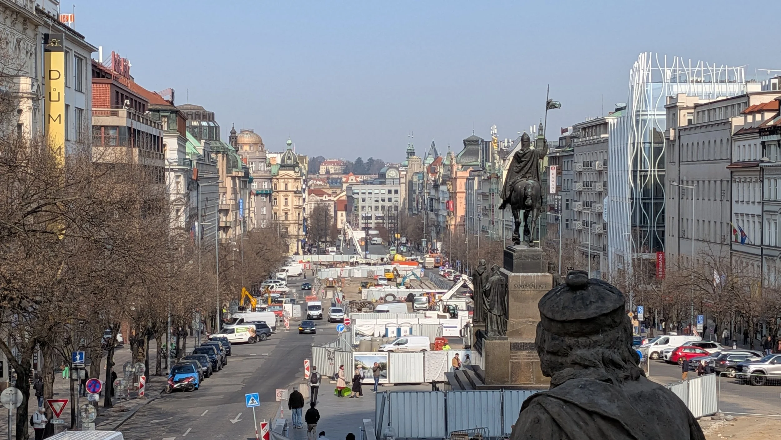 A city street with historical buildings, a statue of a mounted figure, and ongoing construction with vehicles and pedestrians.