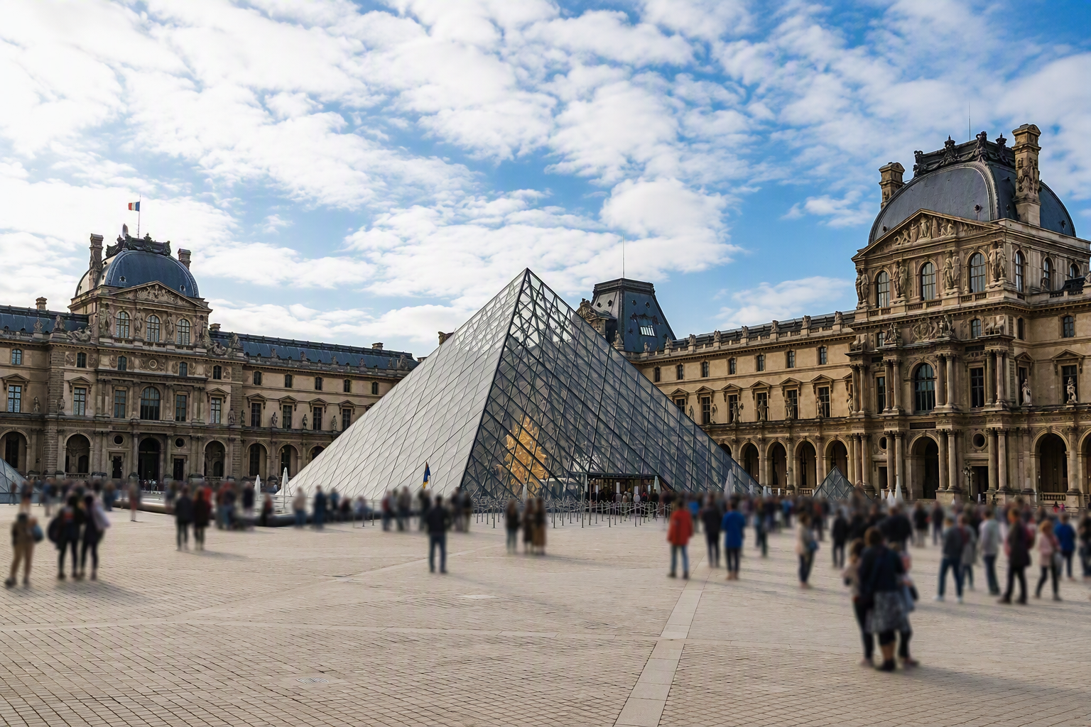 The Louvre Museum in Paris, France, with its glass pyramid entrance and historic palace buildings, under a partly cloudy sky, with numerous visitors walking in the courtyard.