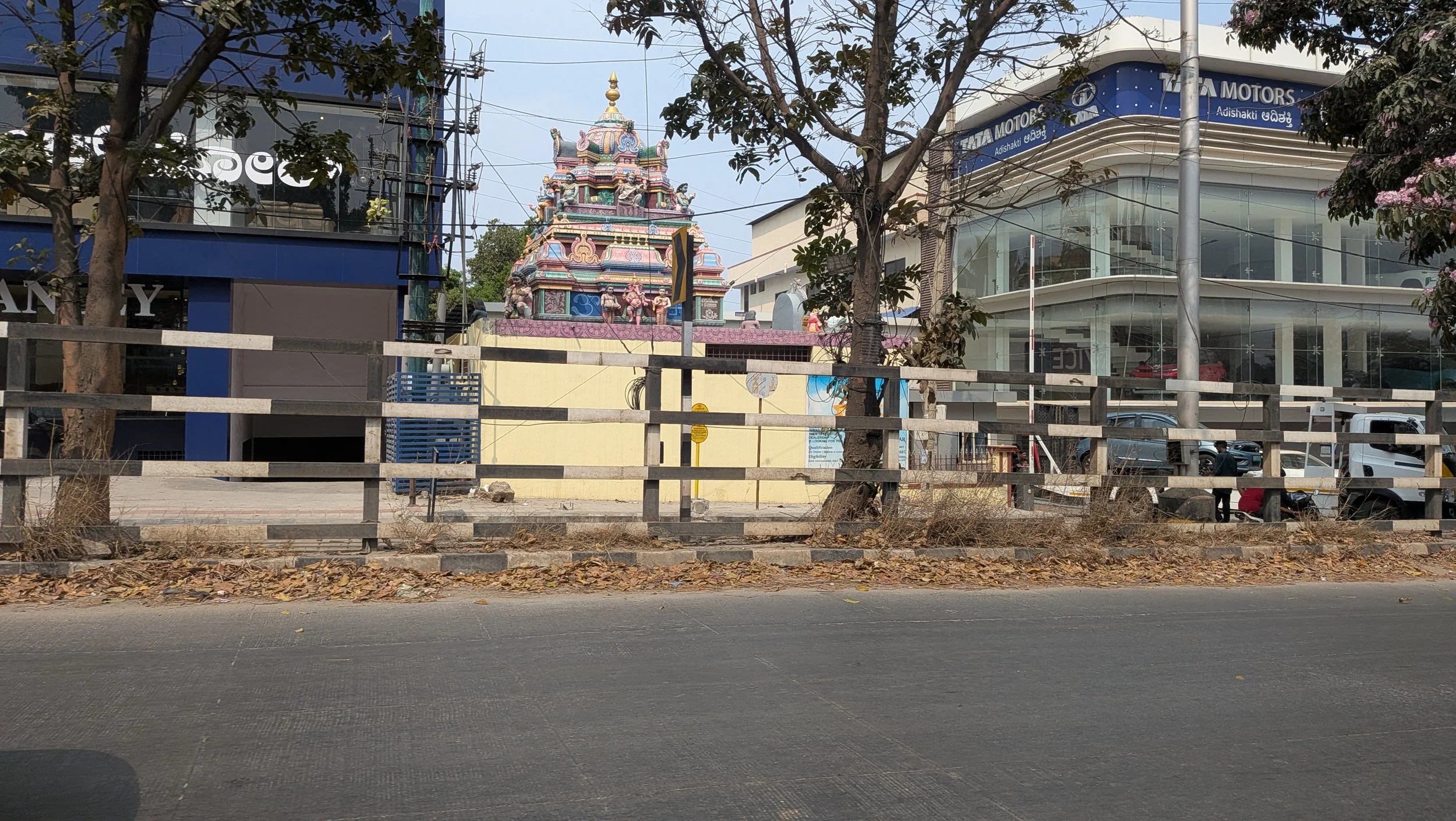 A colorful Hindu temple with a decorated pyramid-shaped tower behind a wooden fence. Modern buildings and parked cars are nearby, and there are trees with leaves and flowers.
