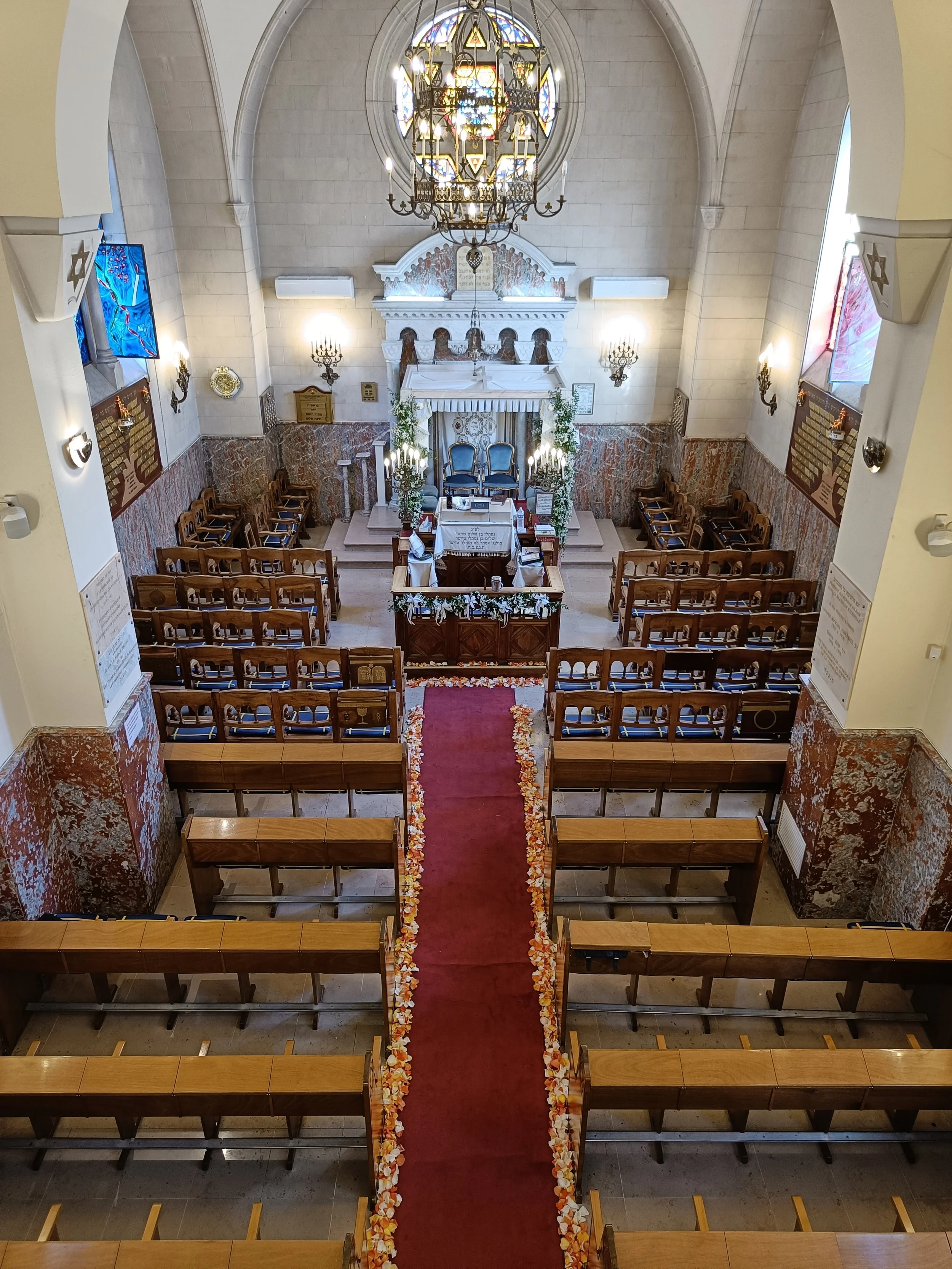 Interior of a church with wooden pews, a red aisle runner decorated with flower petals, an altar with chairs, chandeliers, stained glass windows, and marble walls.