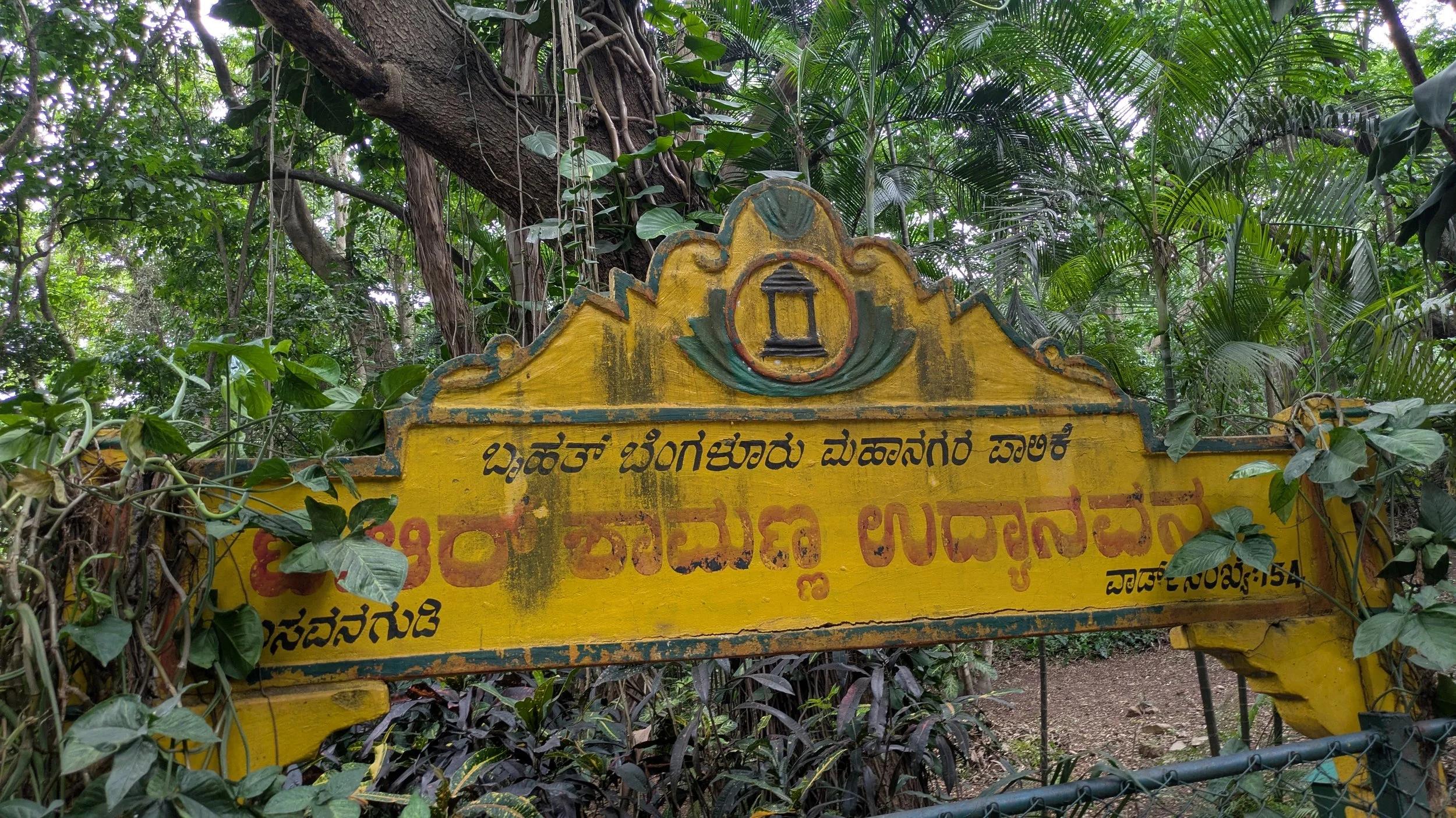 A yellow signboard with Kannada script, surrounded by green foliage and trees in the background.