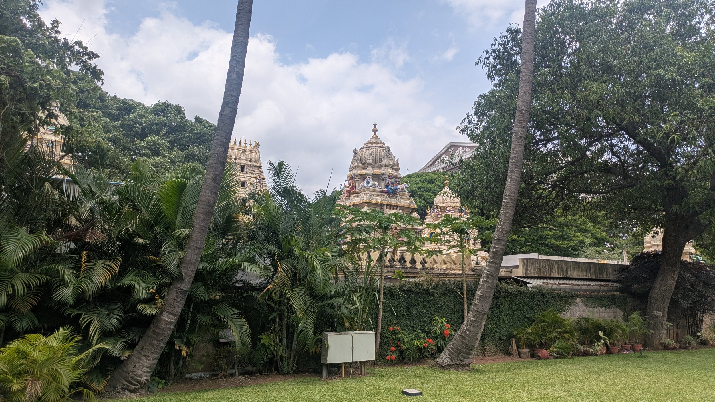 A serene garden scene with lush green plants, palm trees, and vibrant flowers. In the background, ornate Hindu temple architecture with detailed carvings and statues is visible among the trees, under a partly cloudy sky.
