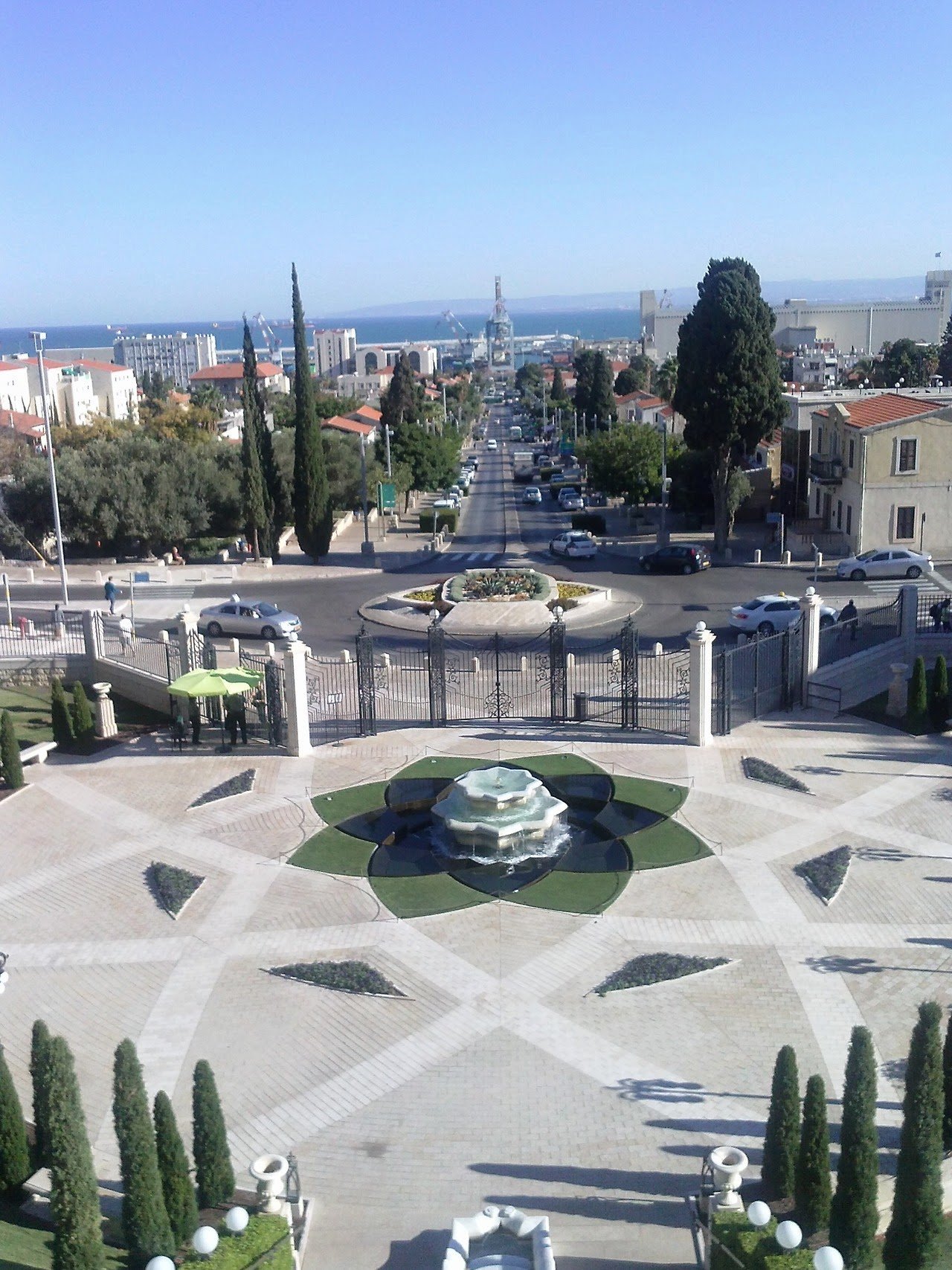 A view of a cityscape with a fountain in the foreground, a park, a street with cars, and industrial cranes near the water in the distance under a clear blue sky.