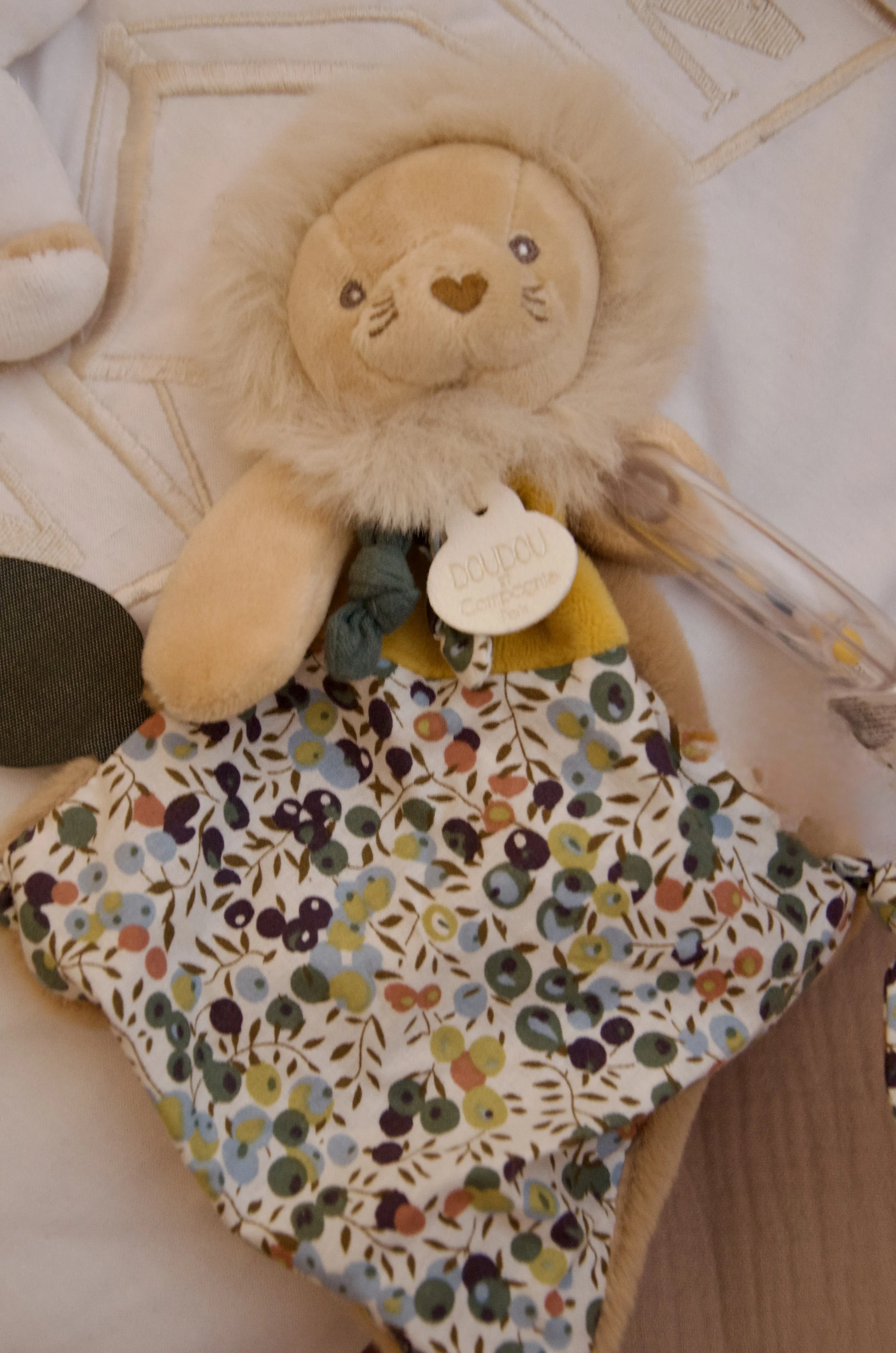 A plush lion toy with a beige mane, embroidered facial features, and a heart-shaped nose. The lion has a tag labeled 'Dauphin Comfort.' It is placed on a bed with a floral bedsheet and other plush toys.