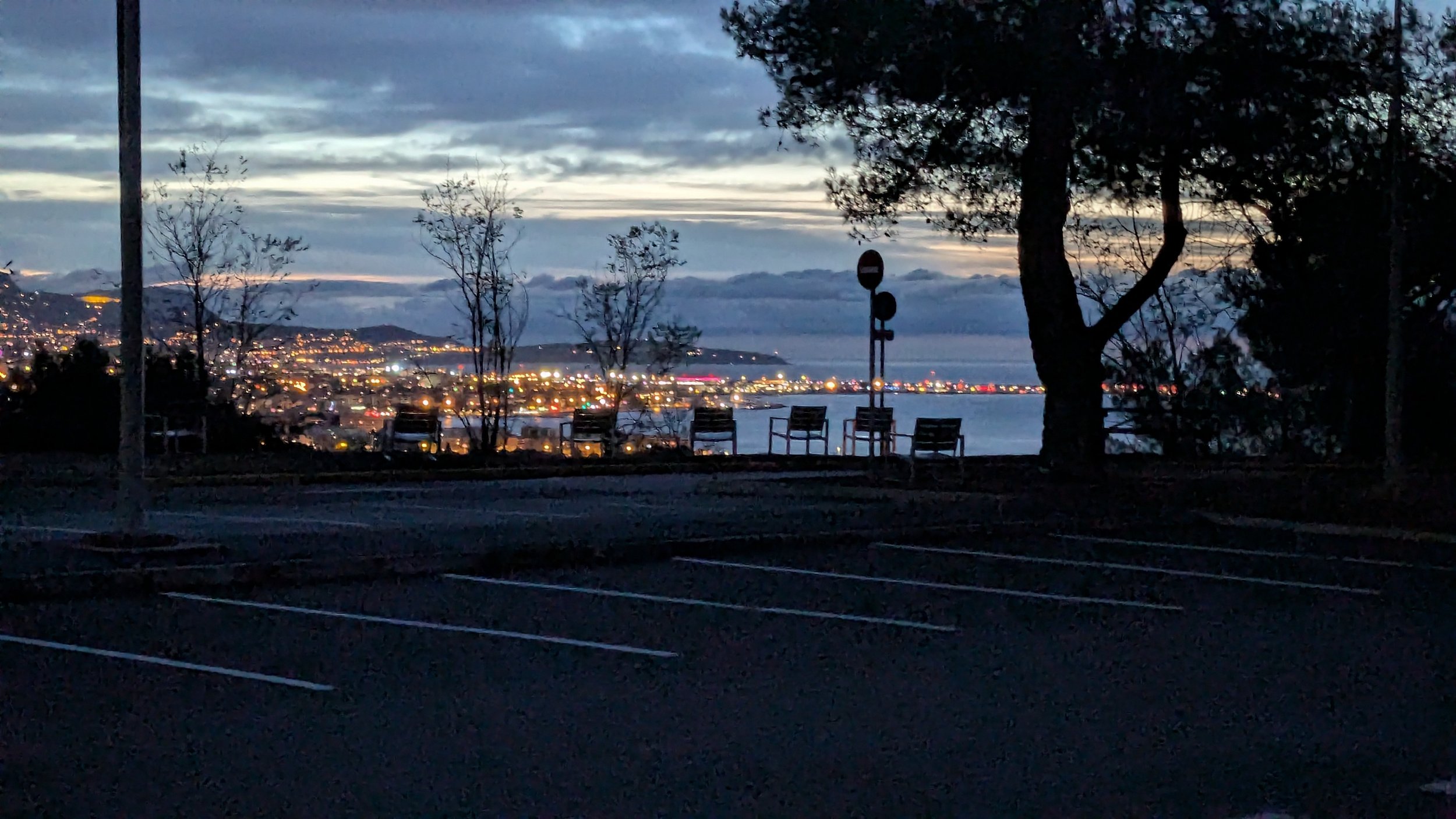 A waterfront scene at dusk with a parking lot in the foreground, leafless trees, and a lakeside city illuminated with lights in the background; cloudy sky overhead.