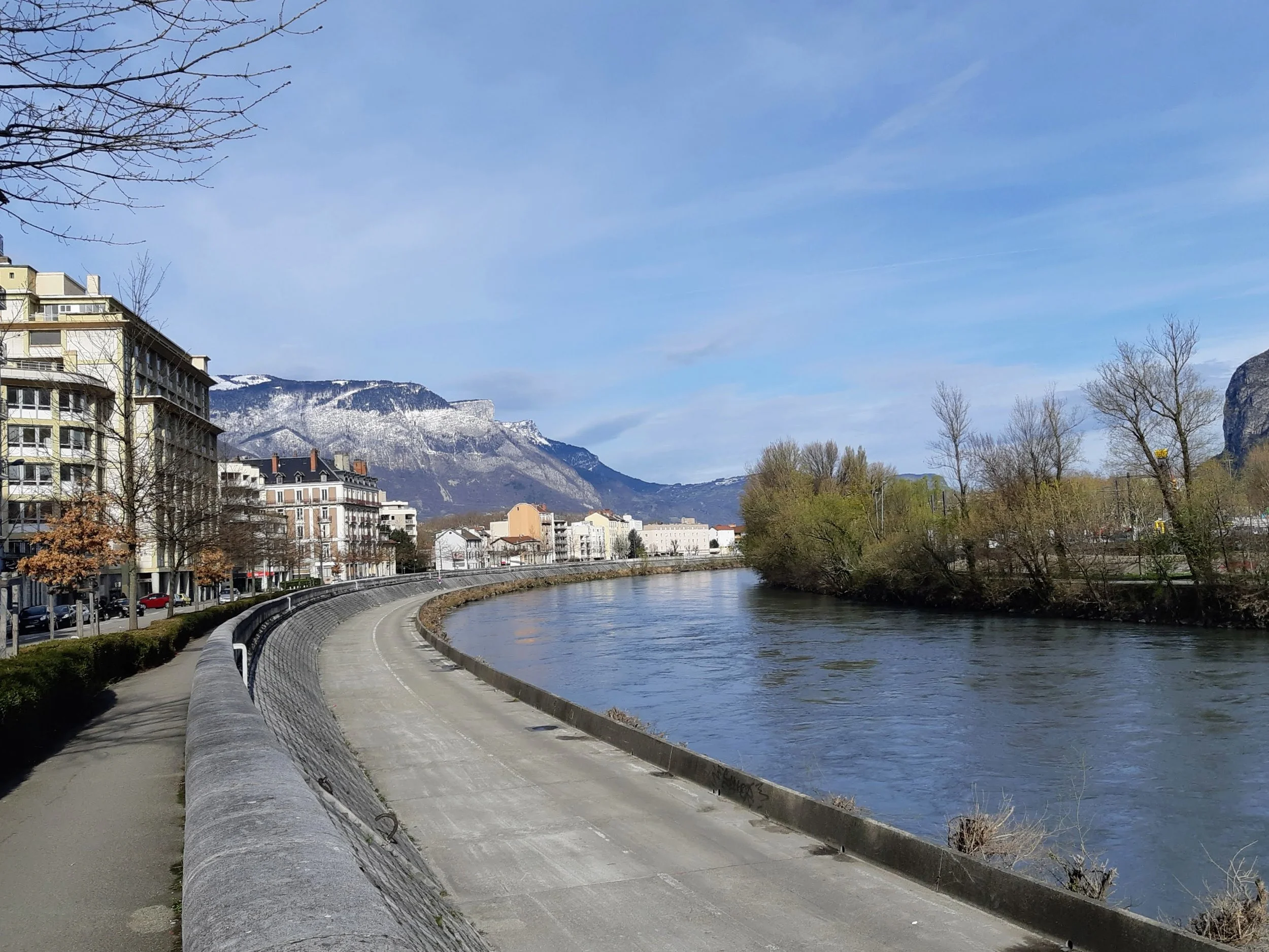 View of a city river promenade with buildings on the left, trees on the right, and snow-capped mountains in the background under a partly cloudy sky.
